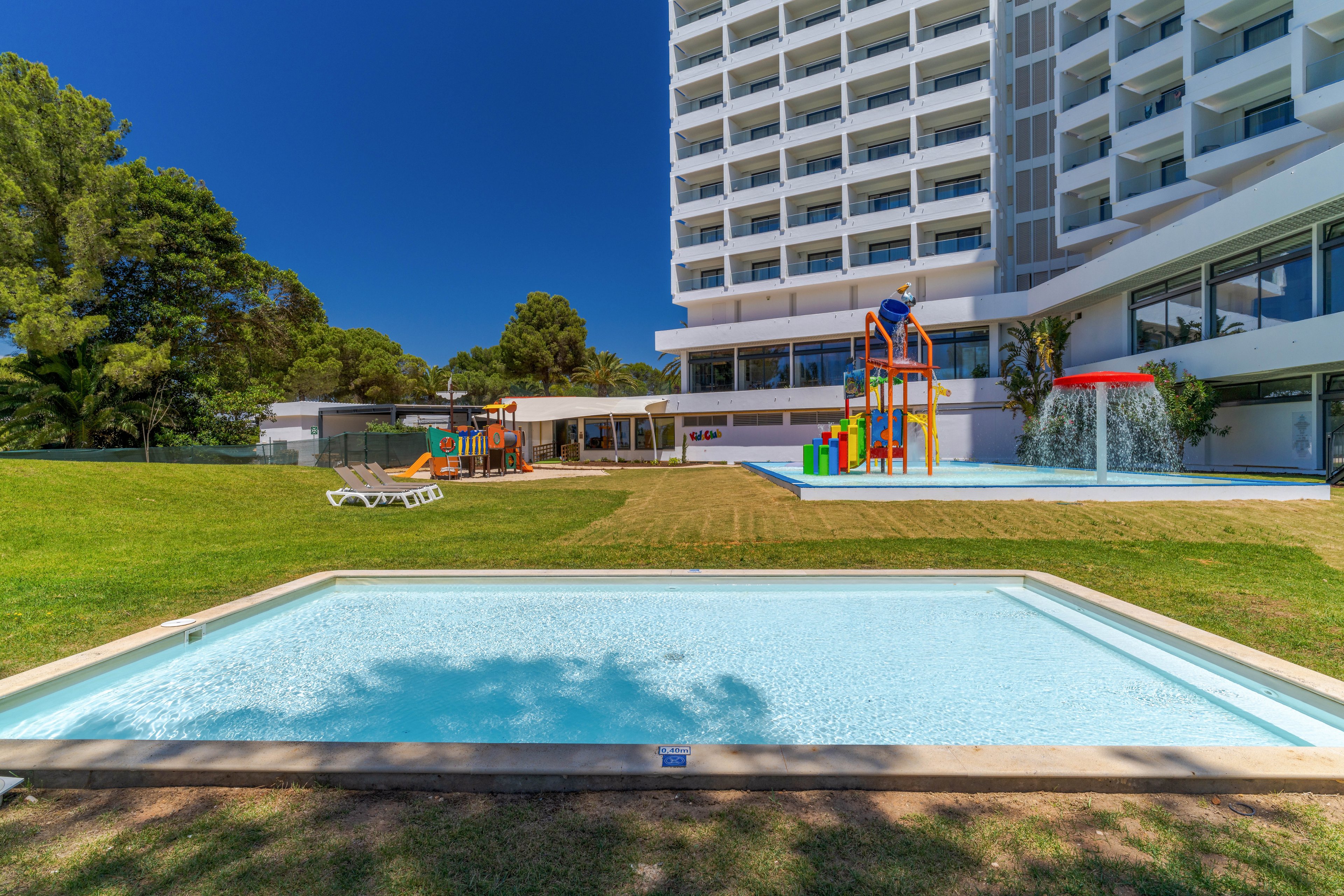 An outdoor hotel recreation area features a shallow rectangular pool in the foreground on a bright sunny day. In the background, a larger children’s splash pool includes colorful slides and a mushroom-shaped water feature. A modern multi-story hotel building rises to the right, with balconies overlooking the green lawn and pool area. Tall trees and clear blue sky frame the relaxing family-friendly resort setting.