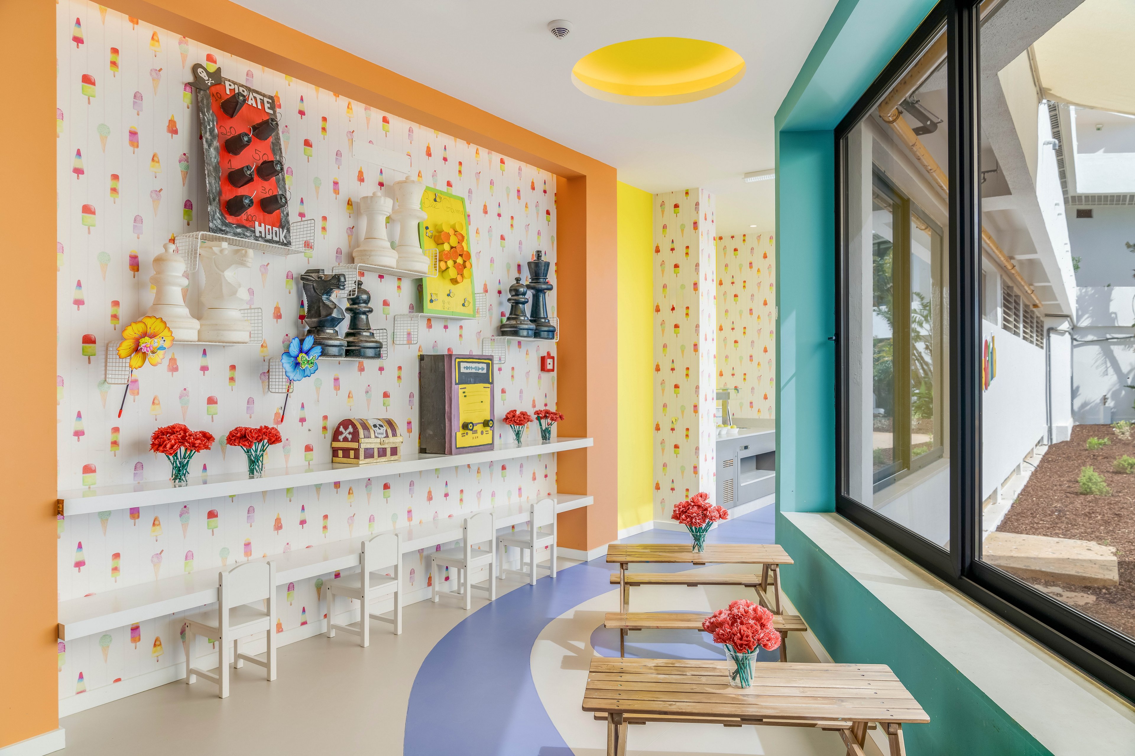 Brightly colored children’s playroom features small wooden tables with flower vases and low white chairs along the wall. The walls are painted in orange, yellow, and teal tones with playful patterned wallpaper and whimsical art pieces. Large chess-piece sculptures, toy houses, and colorful pinwheels decorate a long wall shelf. A curved floor design and large window bring light and movement into the cheerful space.