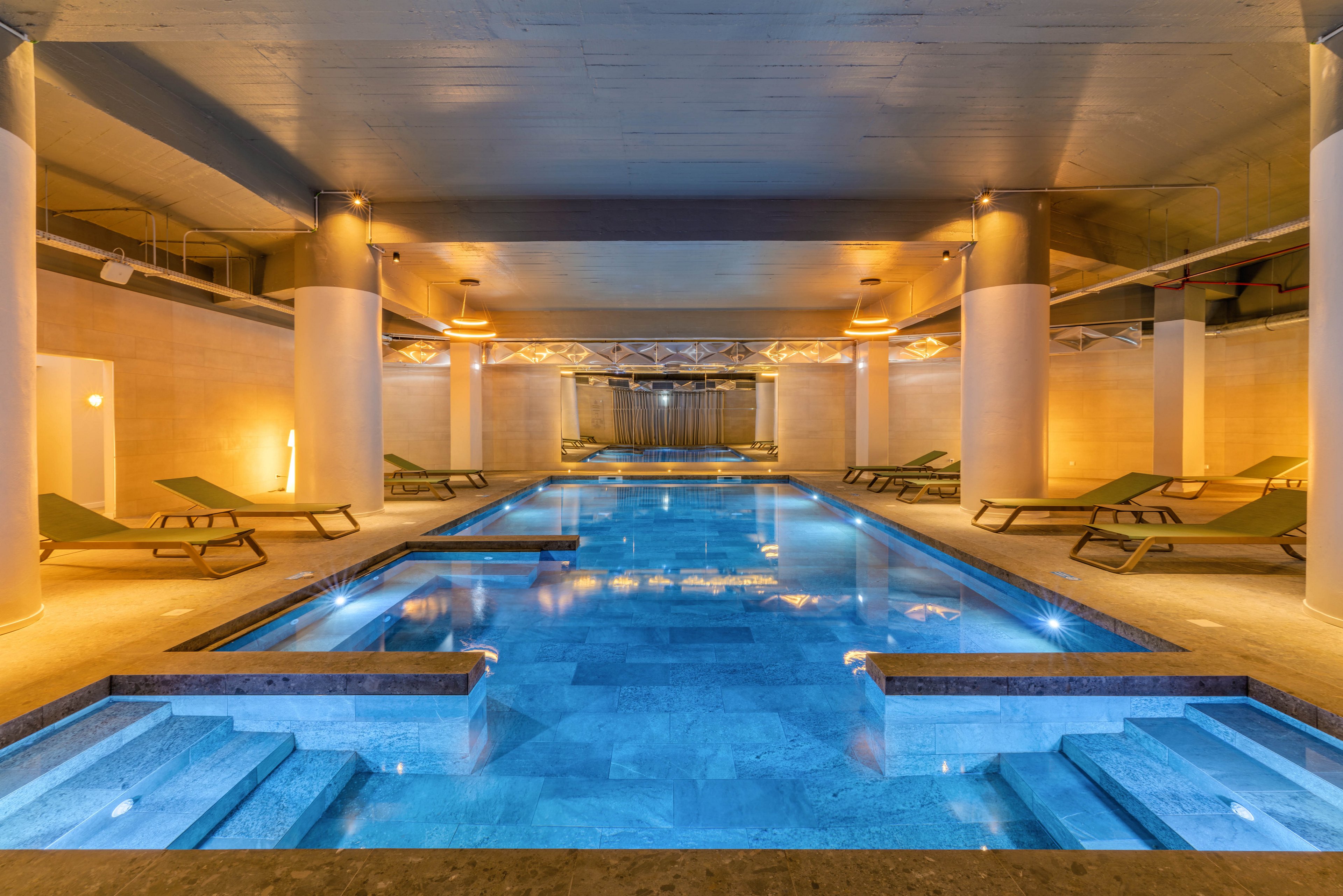A luxurious indoor swimming pool glows with blue lighting in a modern spa setting. Green reclining loungers line both sides of the pool, inviting guests to relax. Warm ceiling and wall lights create a calm contrast to the cool water. The space feels tranquil, symmetrical, and designed for wellness and leisure.