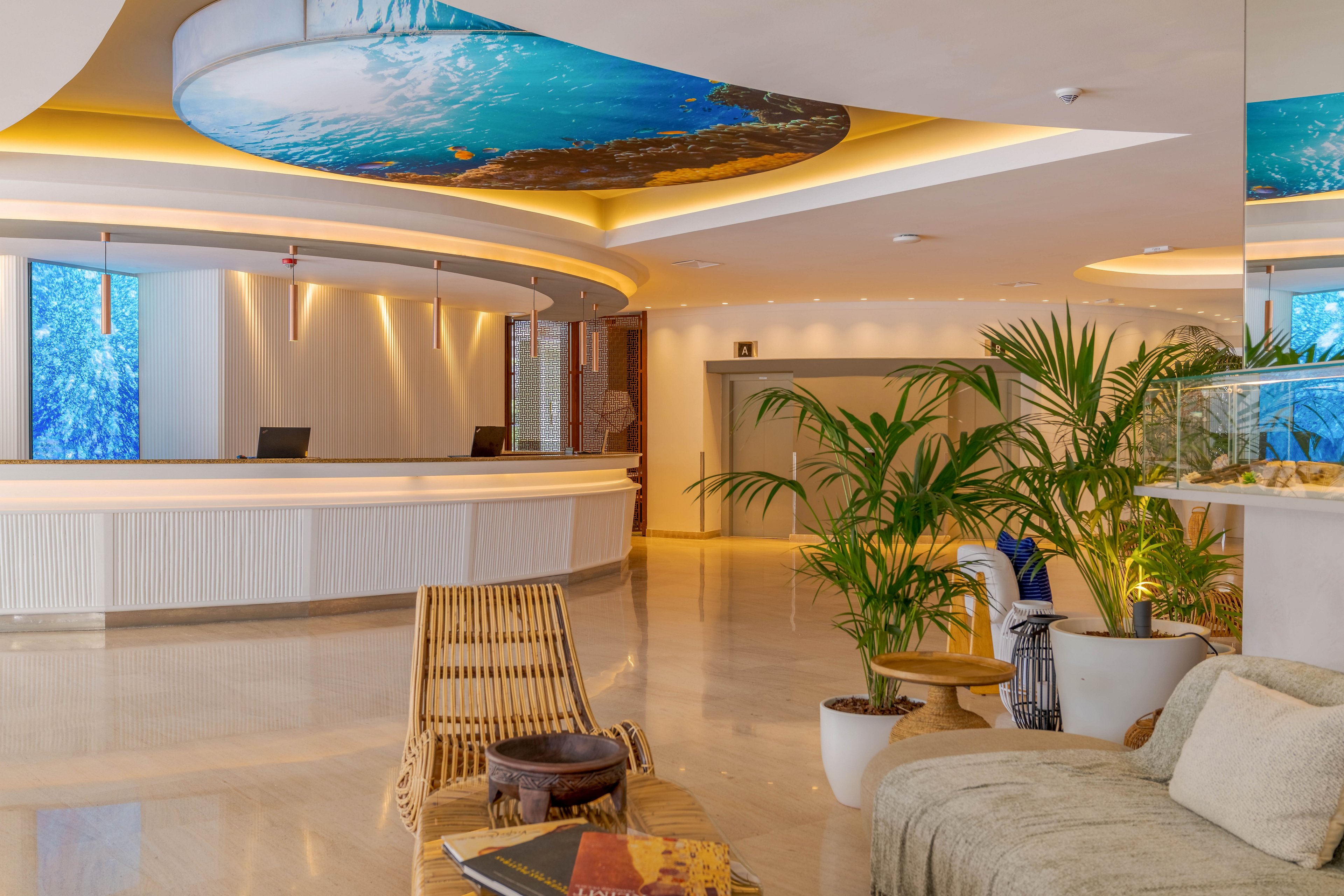A contemporary hotel lobby features a large curved white reception desk in the background. The space is illuminated with warm recessed lighting and blue digital panels behind the counter. In the foreground, comfortable seating, a wicker chair, and potted tropical plants create a relaxed lounge area. A decorative ceiling panel with an ocean-inspired image adds a vibrant focal point to the clean, polished interior.