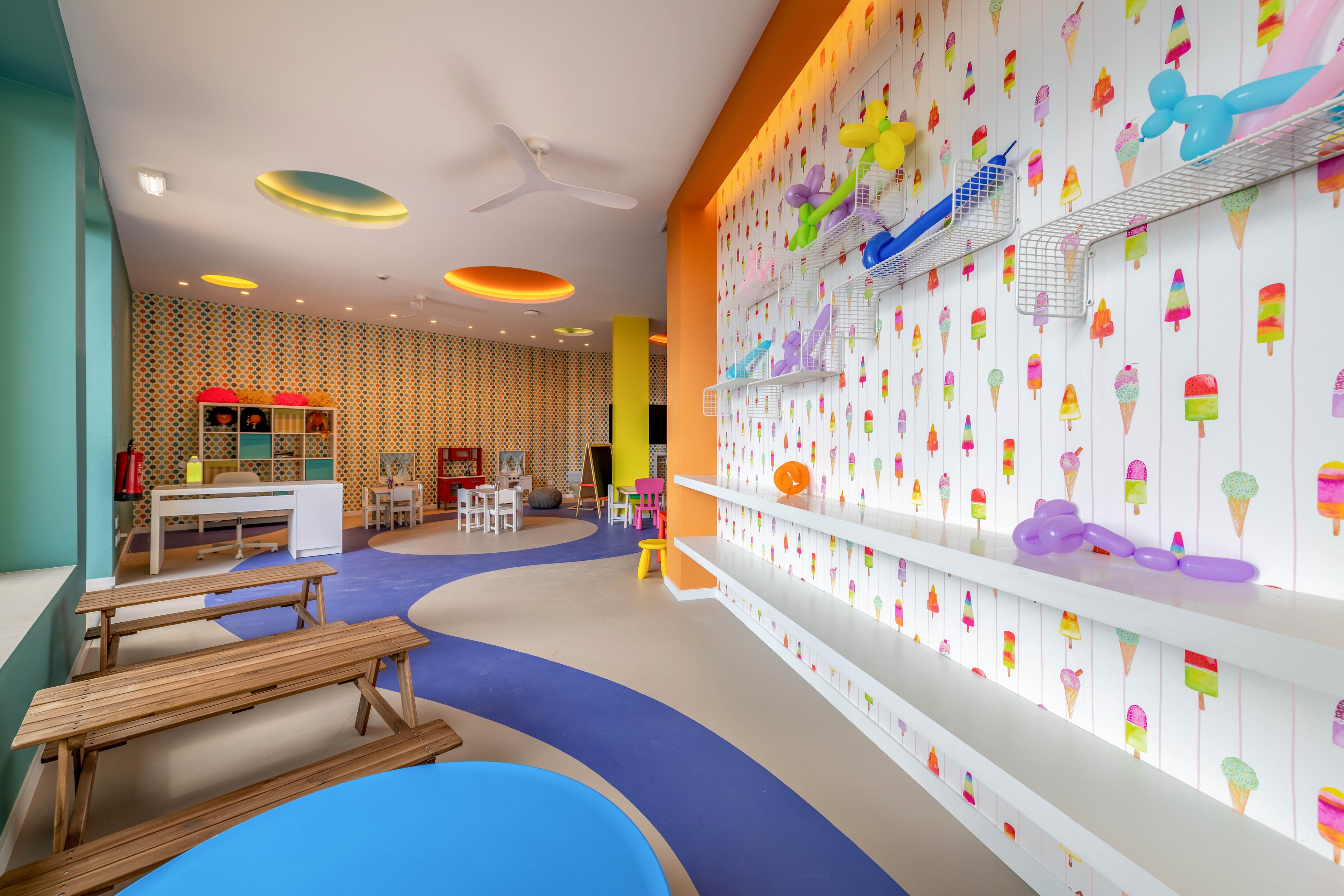 Brightly colored indoor playroom featuring wooden picnic-style benches and curved blue flooring. The walls are decorated with playful ice cream illustrations and balloon animal toys on white shelves. In the background, small tables and chairs create a space for children’s activities and crafts. Warm lighting and bold accent colors give the room a cheerful, inviting atmosphere.