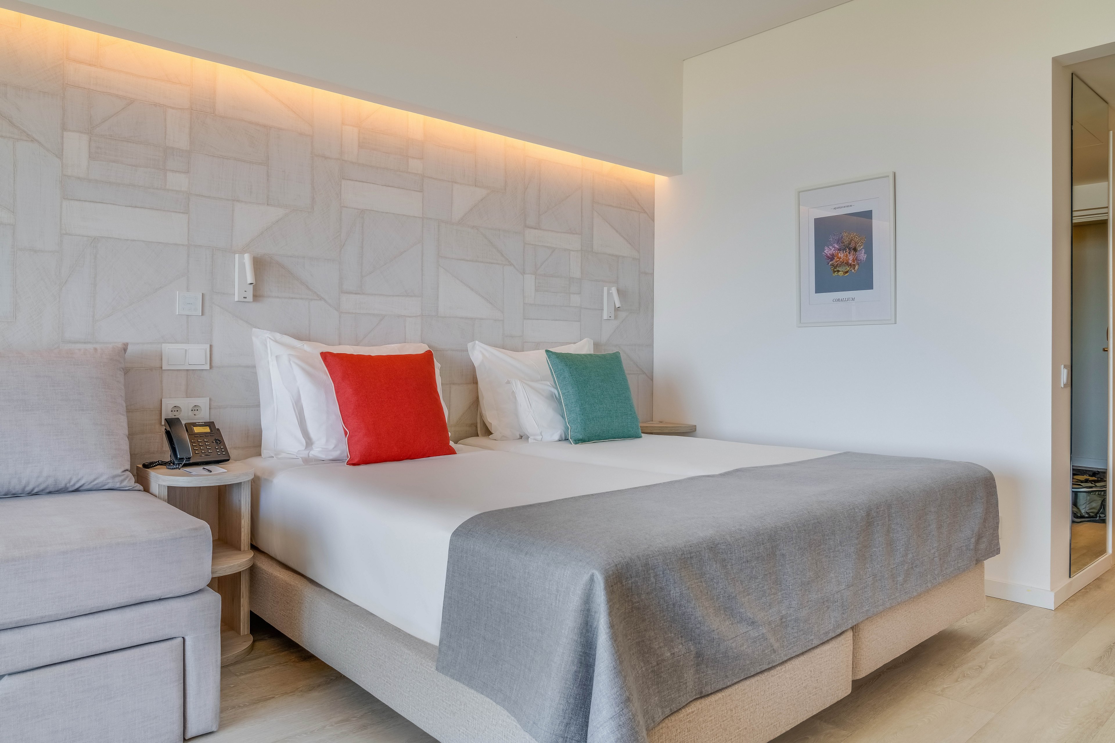 A contemporary hotel bedroom features a neatly made double bed with a light gray bedspread. Bright red and green accent cushions add color against crisp white pillows. A sofa, bedside table with telephone, and wall-mounted reading lights complete the functional space. Soft indirect lighting and a framed artwork on the wall create a calm, inviting atmosphere.