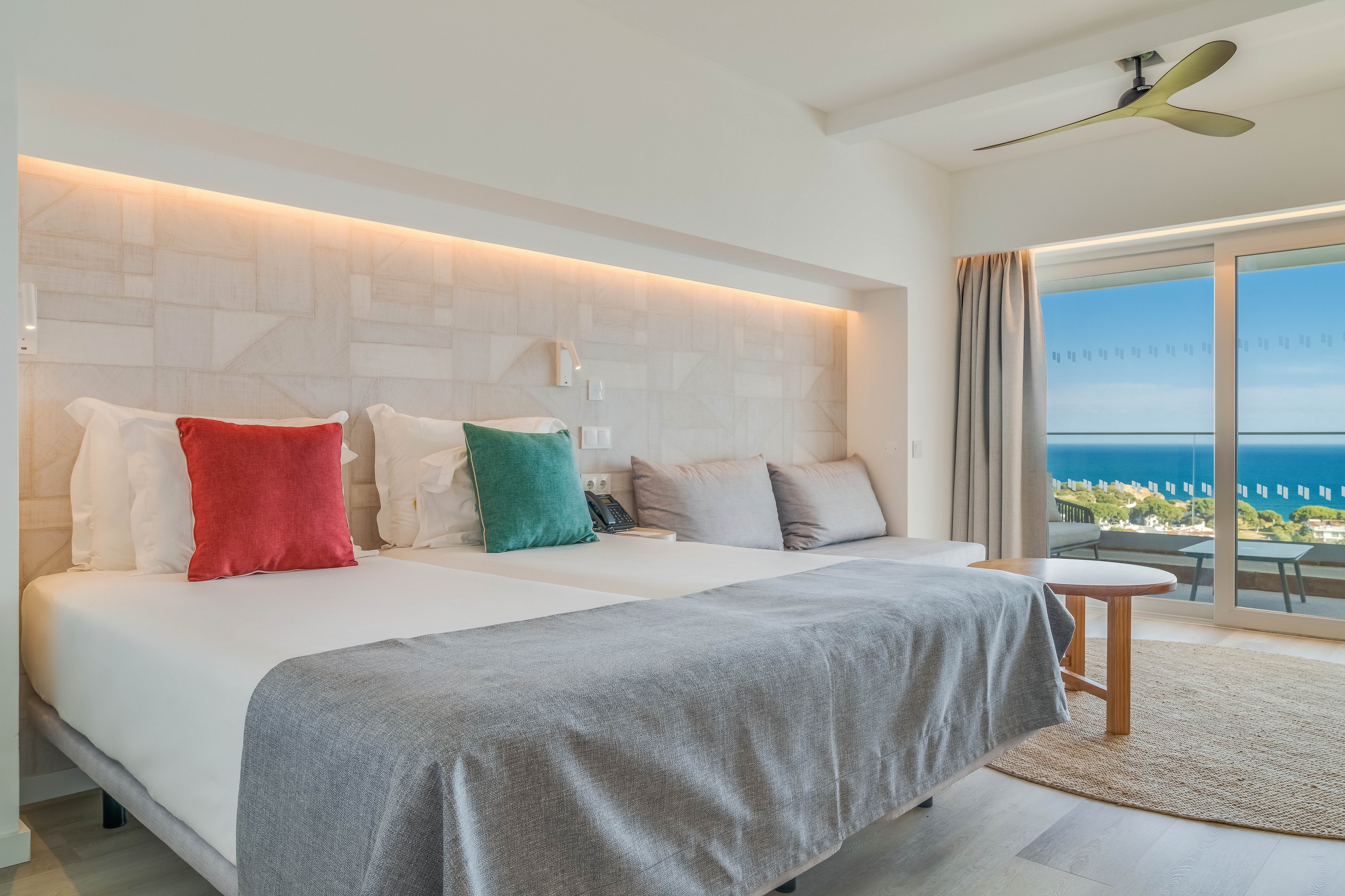 A bright, modern hotel room features twin beds pushed together with white linens and colorful accent pillows. Soft indirect lighting highlights the textured wall and minimalist bedside fixtures. A small seating area with a round table and chairs sits near the sliding glass doors. Beyond the balcony, a clear blue ocean and coastline are visible under a sunny sky, creating a relaxing vacation atmosphere.