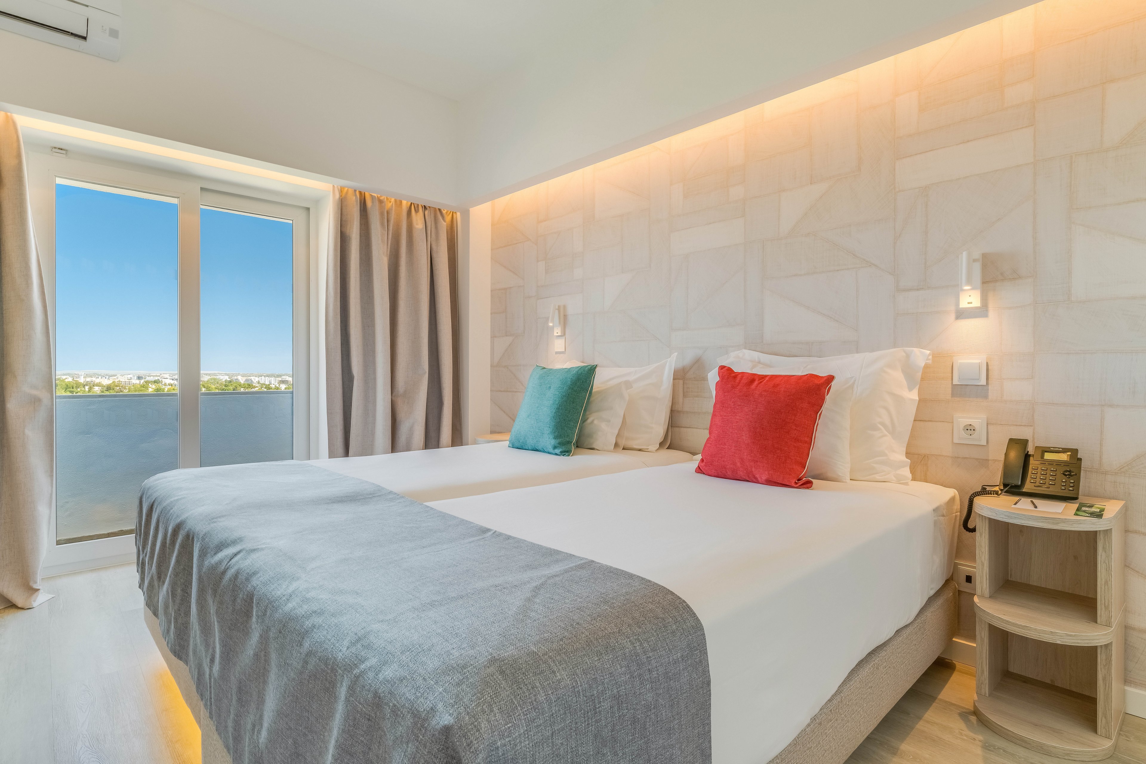 Modern hotel room featuring two neatly made twin beds with light gray throws and colorful accent pillows. The space is bright and airy, with large glass doors leading to a balcony and a clear blue sky outside. Soft indirect lighting runs along the textured wall behind the beds, creating a warm and inviting atmosphere. A small bedside table holds a telephone and notepad, emphasizing the room’s functional comfort for guests.