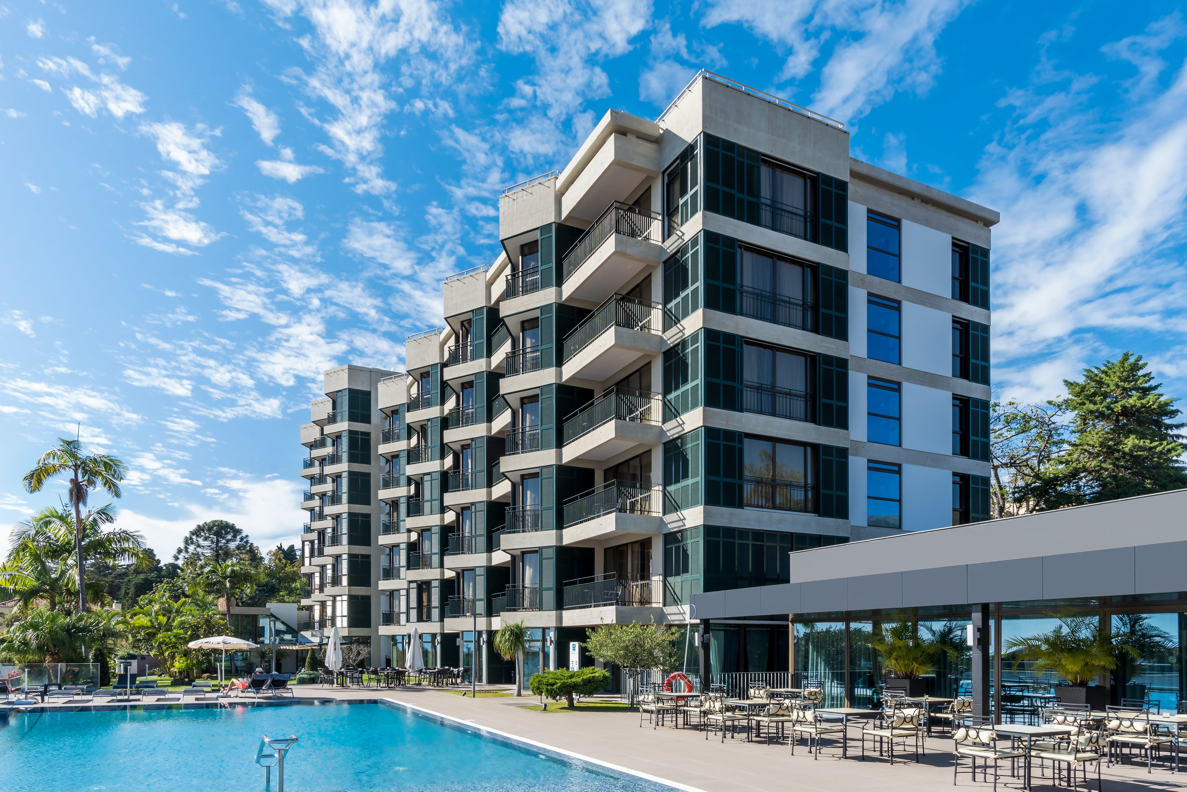 A contemporary multi-story hotel featuring glass balconies and a spacious outdoor pool area. The setting includes palm trees and clear blue skies, creating a relaxing atmosphere. Outdoor seating is arranged near the pool, ideal for leisure or dining.