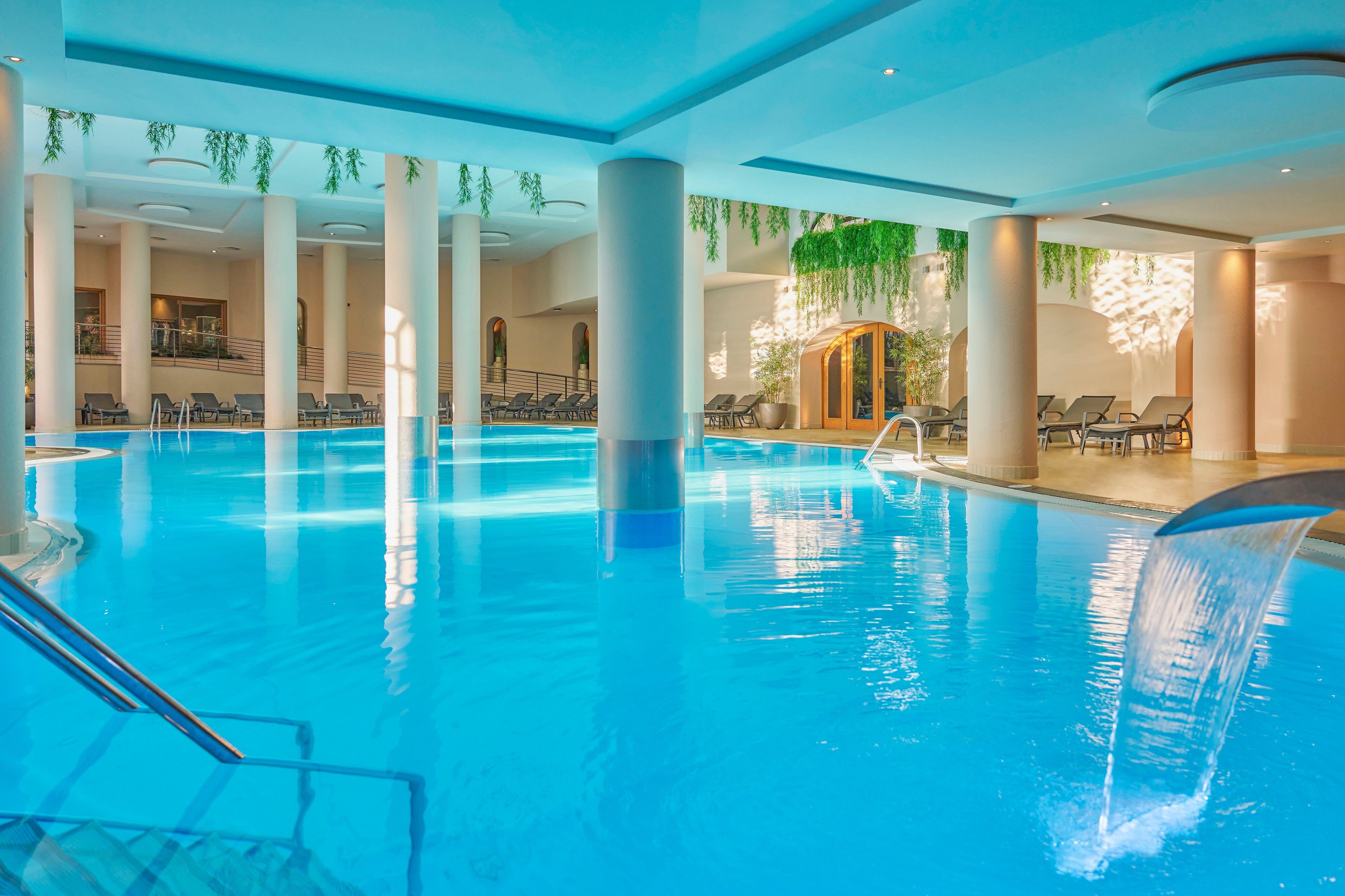 Bright turquoise water fills a spacious indoor spa pool surrounded by white columns. Multiple waterfall features pour into the pool, creating a relaxing and refreshing atmosphere. Lounge chairs line the perimeter, offering seating for guests in a resort-like setting. Hanging greenery and warm ambient lighting add a tranquil, wellness-focused mood.