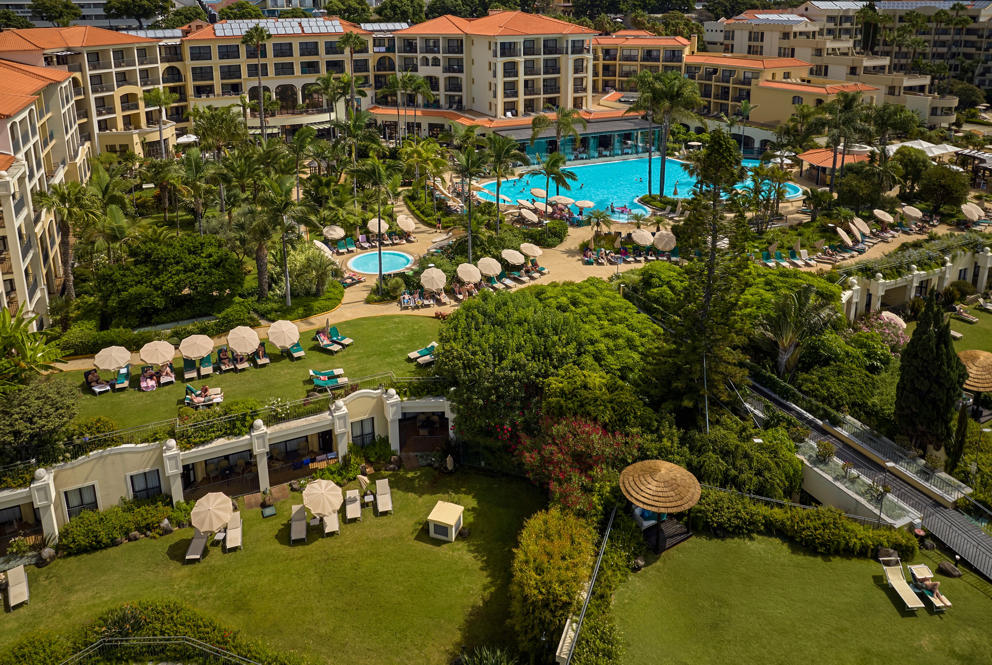 A large resort complex features multiple outdoor swimming pools surrounded by sun loungers and umbrellas. The scene is captured from above, showing manicured lawns, lush gardens, and palm trees throughout the property. Guests relax on deck chairs near the pools and in private terrace areas, suggesting a warm, sunny vacation atmosphere. Multi-story hotel buildings frame the central pool area, emphasizing the scale of the destination.