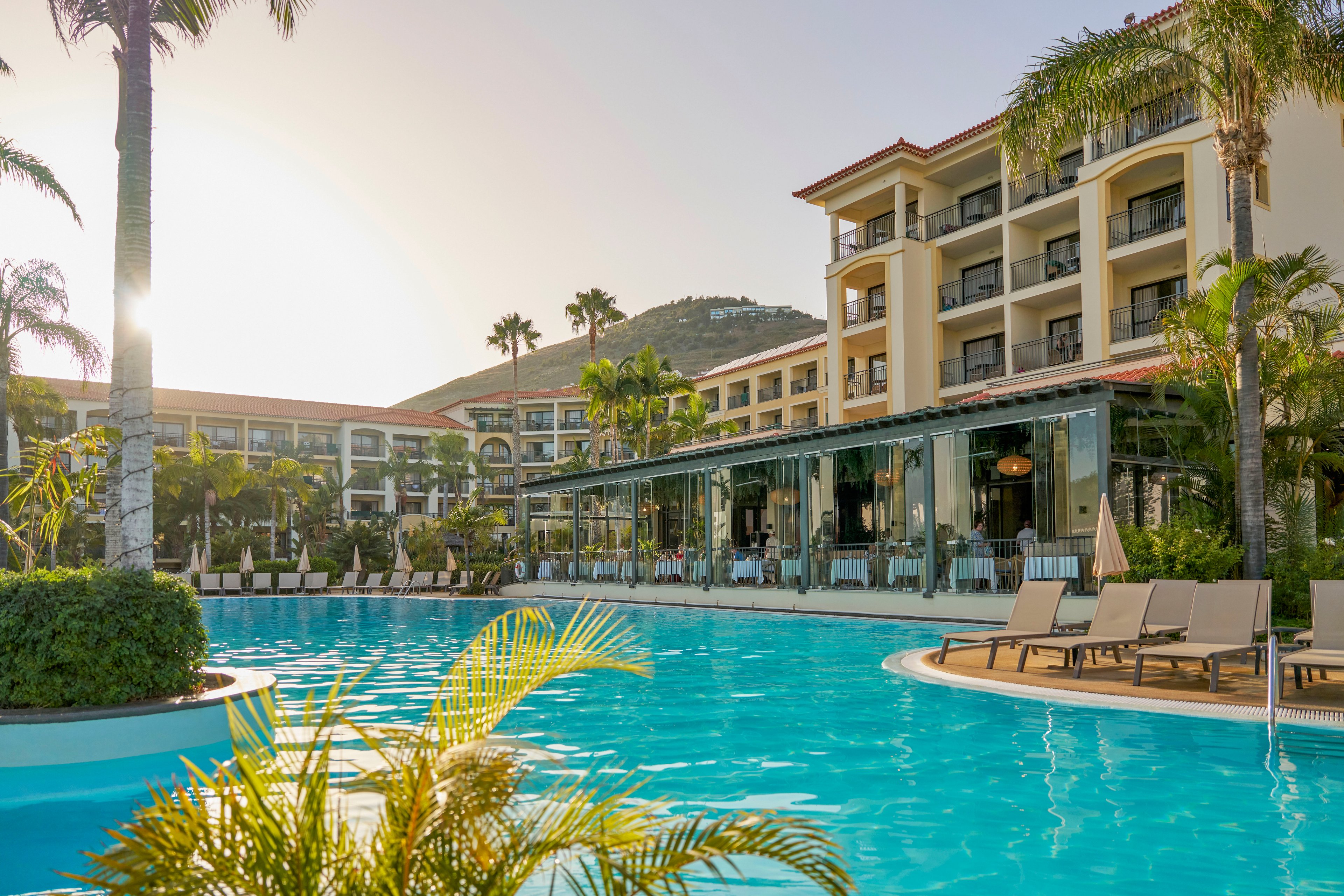 A large outdoor swimming pool sits in front of a multi-story resort building surrounded by lush palm trees. Lounge chairs line the poolside, suggesting a relaxing vacation setting. The sun is low in the sky, casting a warm golden light across the water and buildings. A distant hill rises behind the resort, enhancing the tropical, tranquil atmosphere.