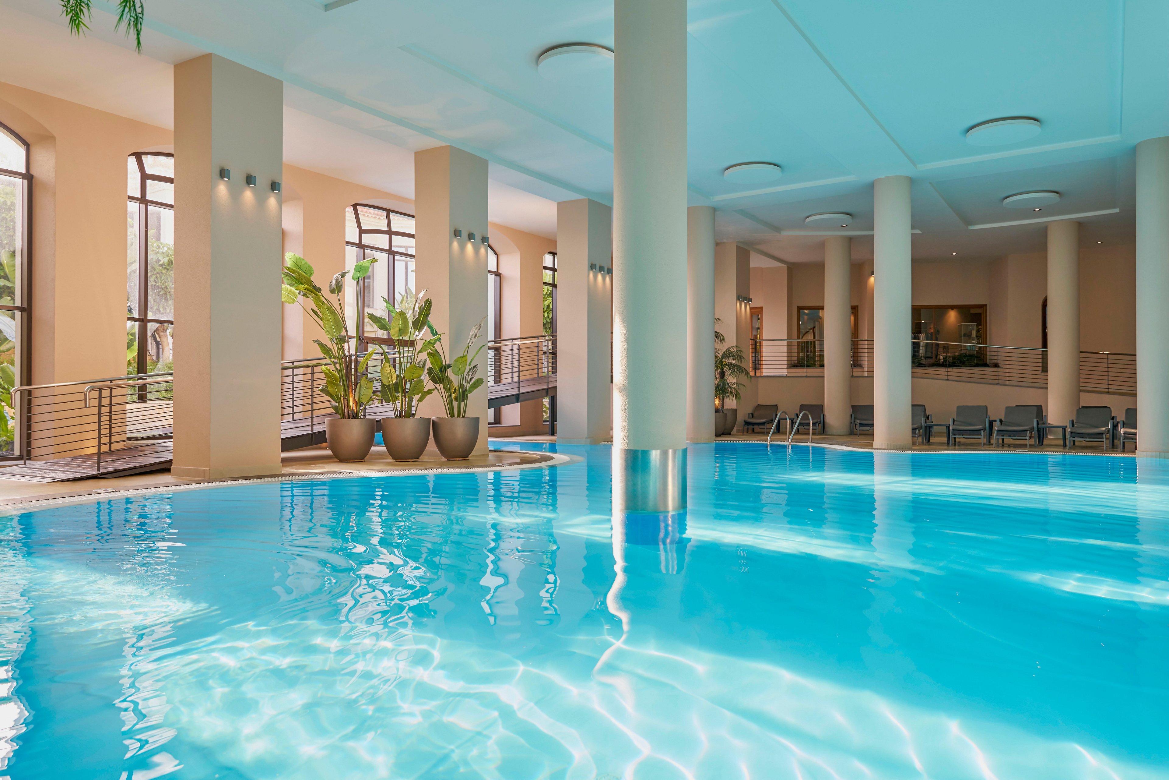 Spacious indoor swimming pool with clear blue water is featured in a modern leisure facility. Tall columns and large arched windows create an open, airy atmosphere with natural light. Potted green plants line the edge of the pool, adding a touch of nature to the contemporary interior. Lounge chairs and a metal handrail suggest a relaxing space for guests to rest and enjoy the poolside environment.