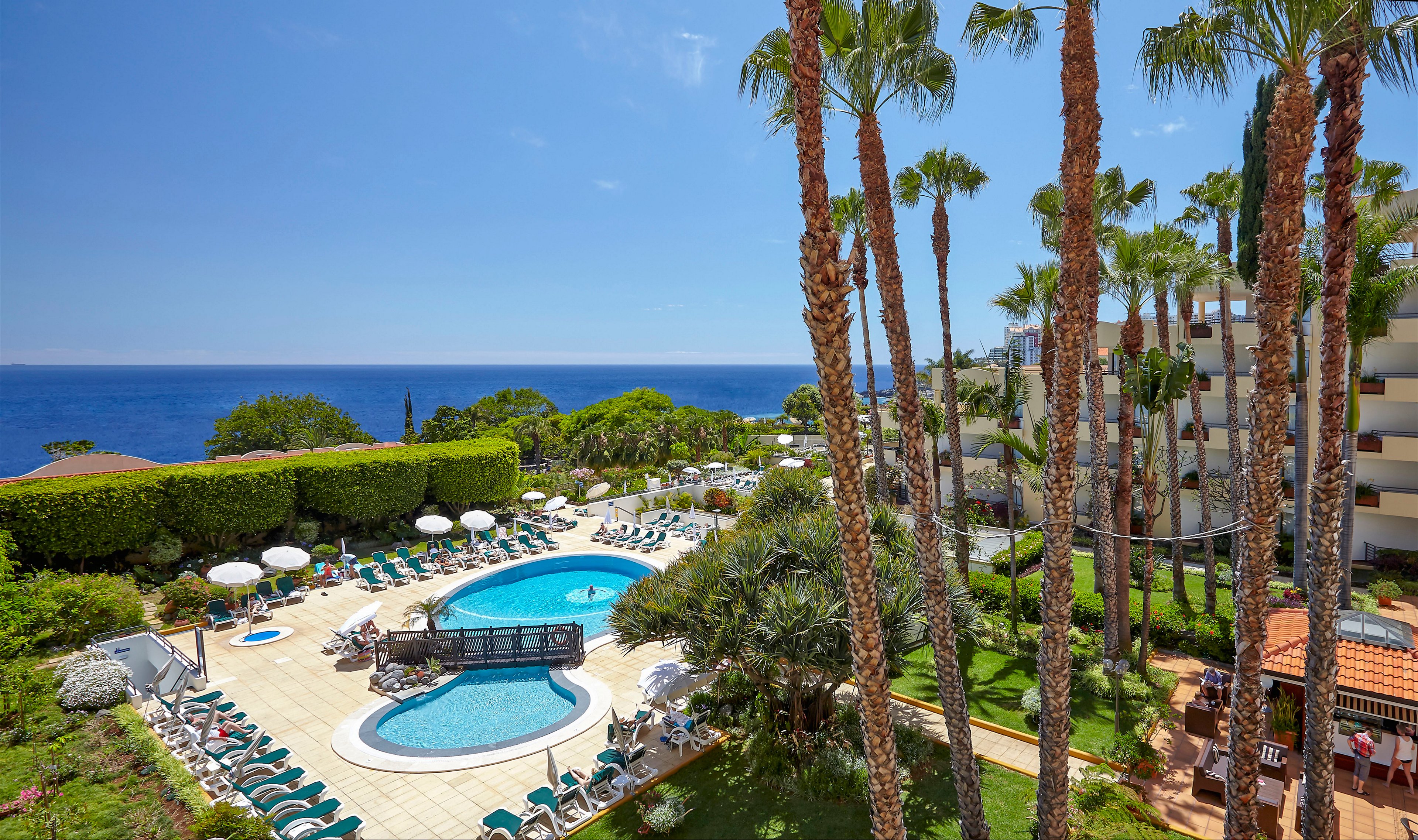 A bright seaside resort features a central outdoor swimming pool surrounded by sun loungers and umbrellas. Tall palm trees frame the scene, with manicured hedges and gardens adding lush greenery. A multi-story hotel building stands to the right, overlooking the pool area and the expansive ocean in the background. The clear blue sky and calm water create a relaxed vacation atmosphere.