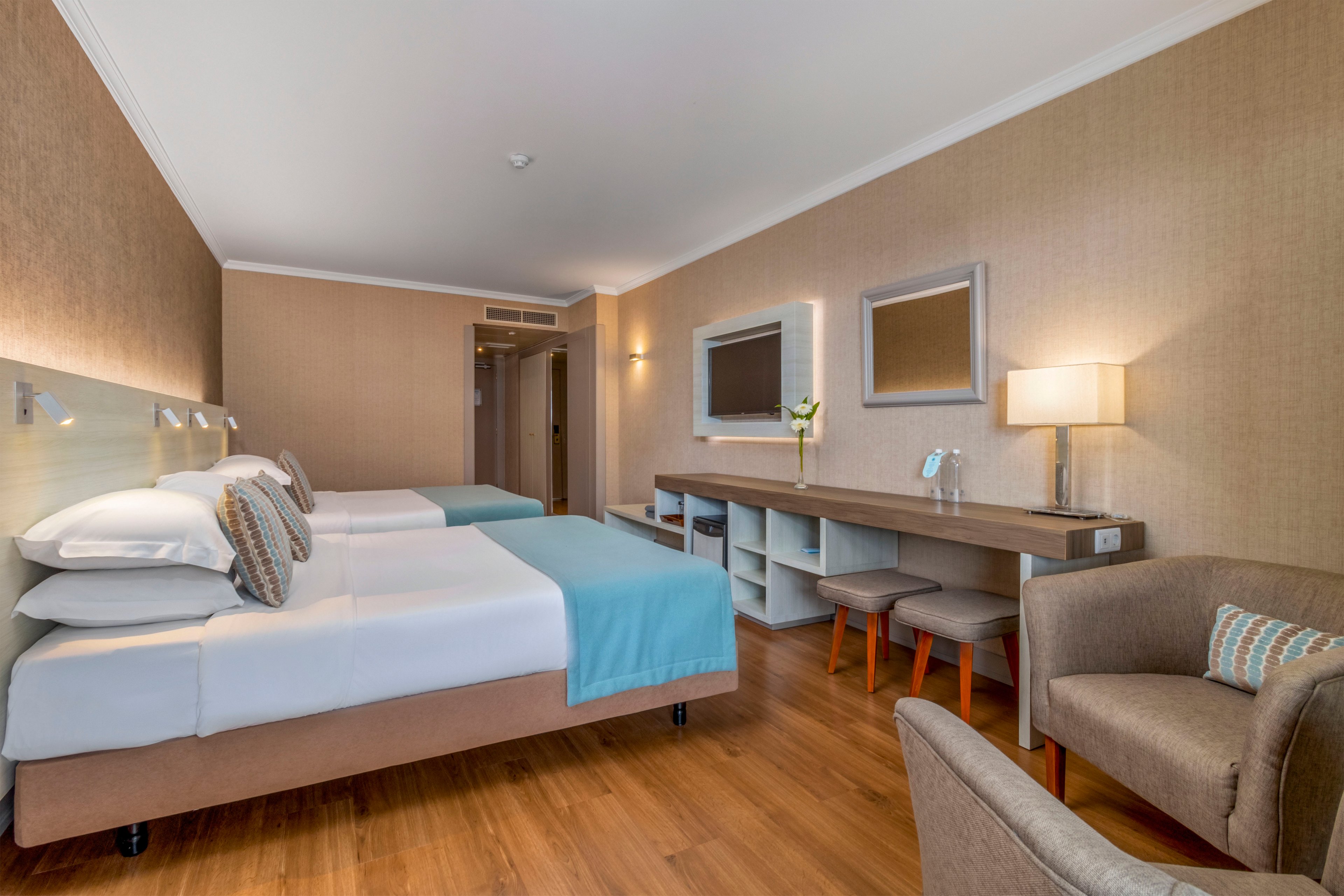A contemporary hotel room features two neatly made twin beds with white linens and light blue throws. The space includes a long desk with a lamp, decorative vase, and two orange stools beneath open shelving. A comfortable armchair with a patterned cushion sits near the beds, and two wall mirrors add depth to the room. Warm beige walls and wooden flooring create a clean, inviting atmosphere under soft ambient lighting.