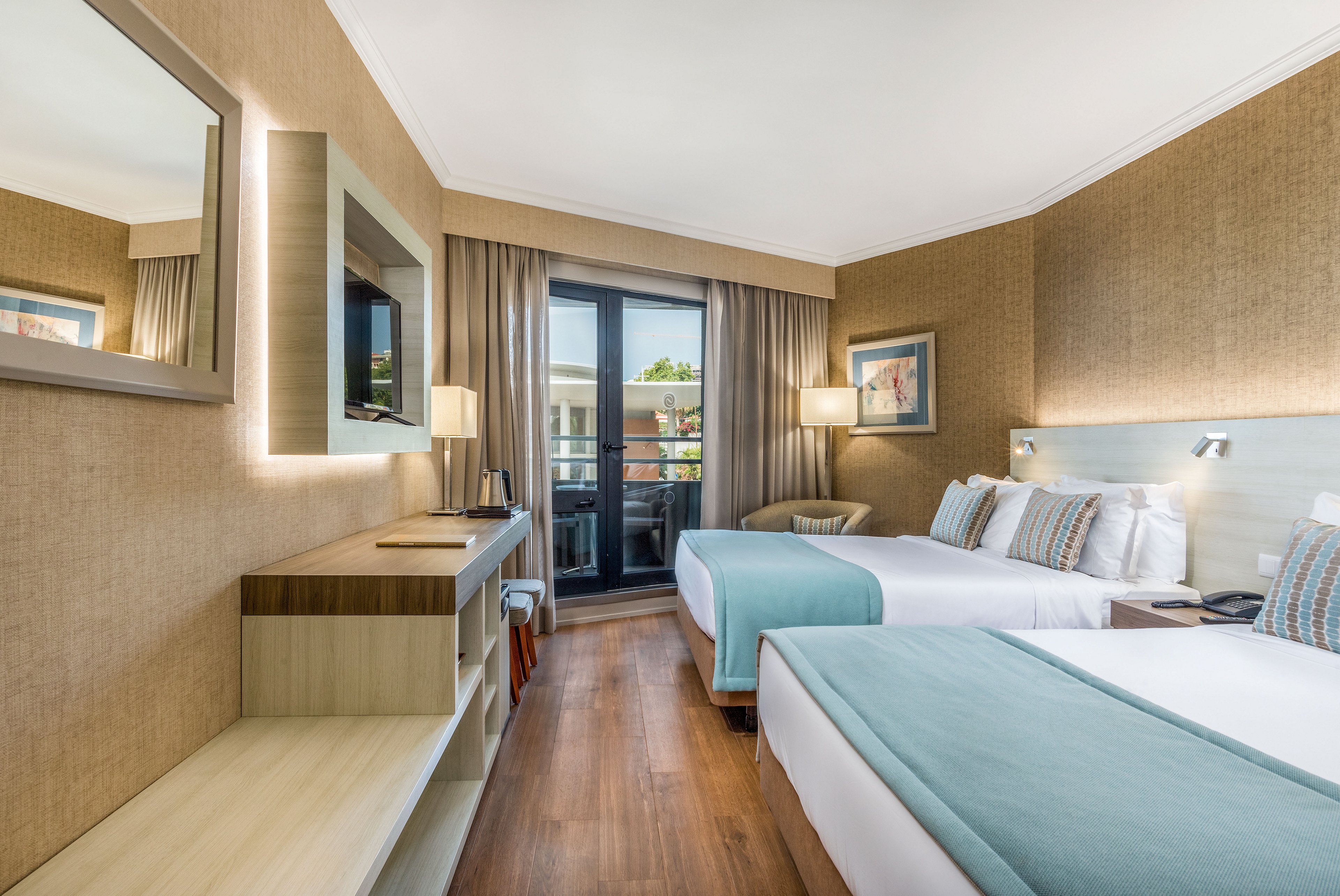 Contemporary hotel guest room featuring two neatly made twin beds with light blue bedding and accent pillows. The space includes a wall-mounted television, desk area, and large mirror along one side. Floor-to-ceiling glass doors open to a balcony with outdoor view, framed by beige curtains. Warm neutral walls and wooden flooring create a clean, inviting atmosphere suitable for leisure or business travel.