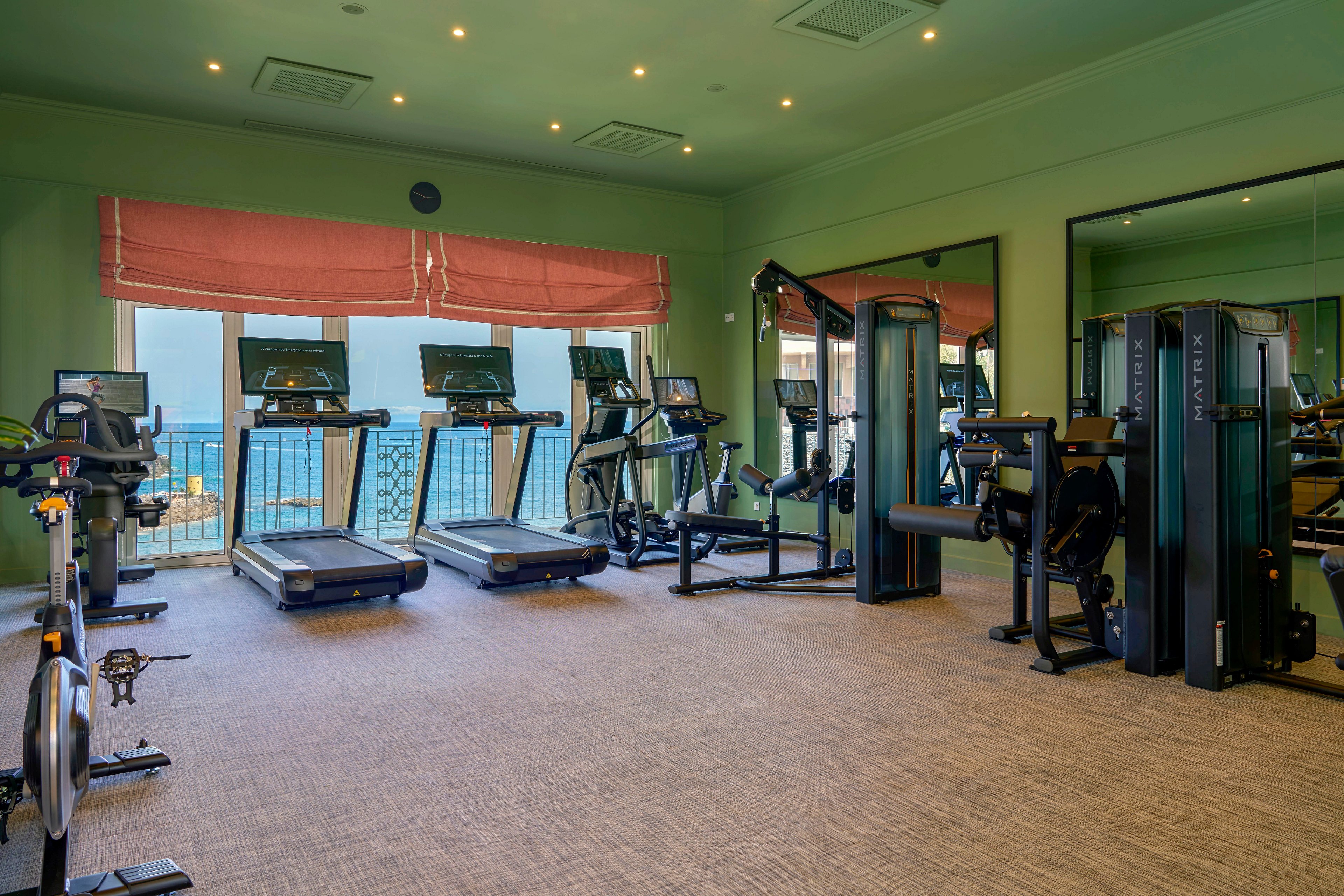 A contemporary indoor gym features multiple treadmills and elliptical machines arranged in front of large windows. Strength-training equipment and weight machines line the opposite side of the room. Soft overhead lighting and muted green walls create a calm, inviting workout environment. The space appears clean, organized, and ready for guests to exercise.