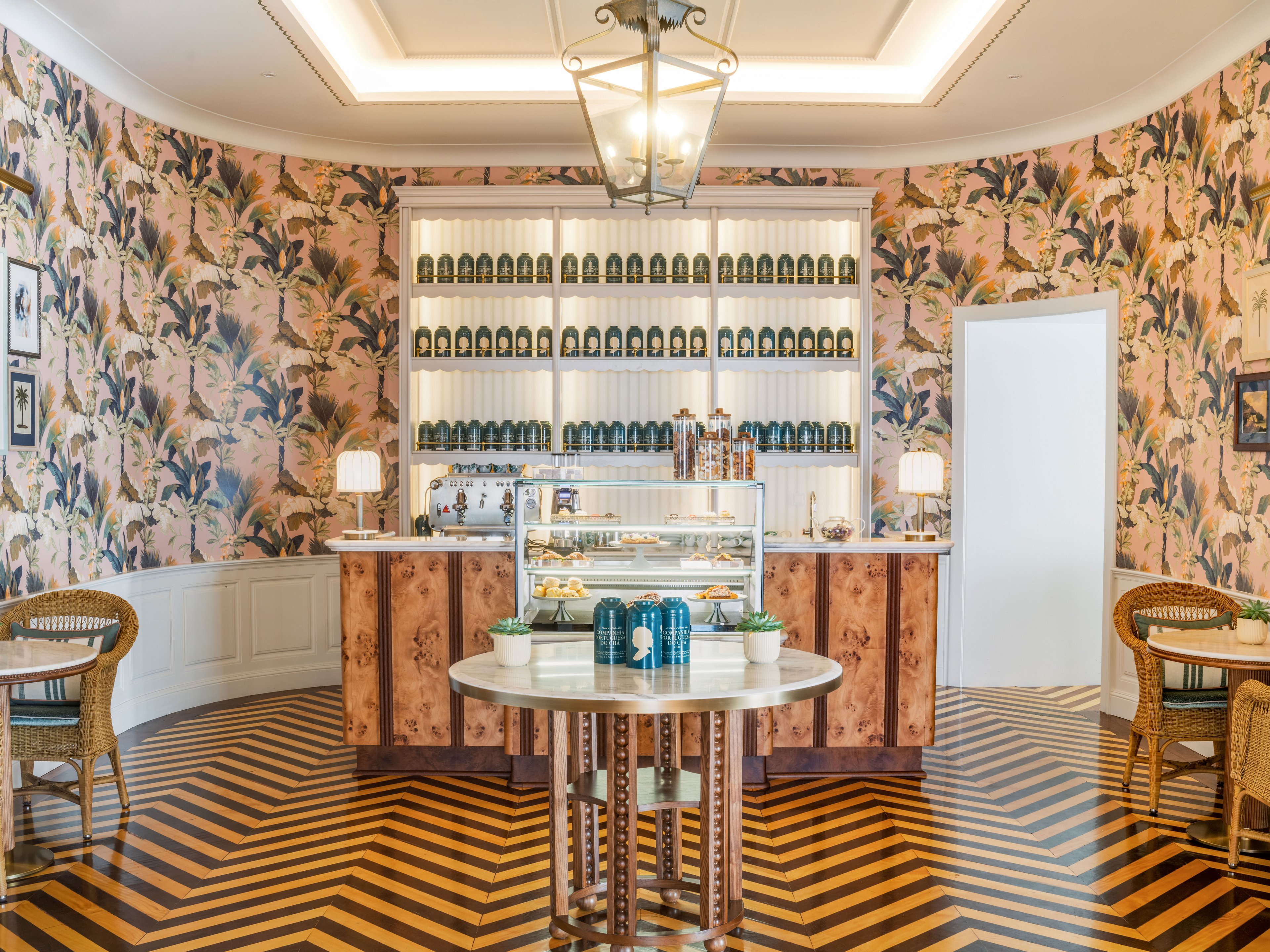A stylish boutique café features a central round table and a wooden counter with a glass display of pastries. The walls are covered in floral wallpaper and lined with shelves of identical green bottles. Two small tables with woven chairs sit on either side, creating an intimate seating area. A geometric chevron wood floor and warm lighting add to the sophisticated, vintage-inspired atmosphere.