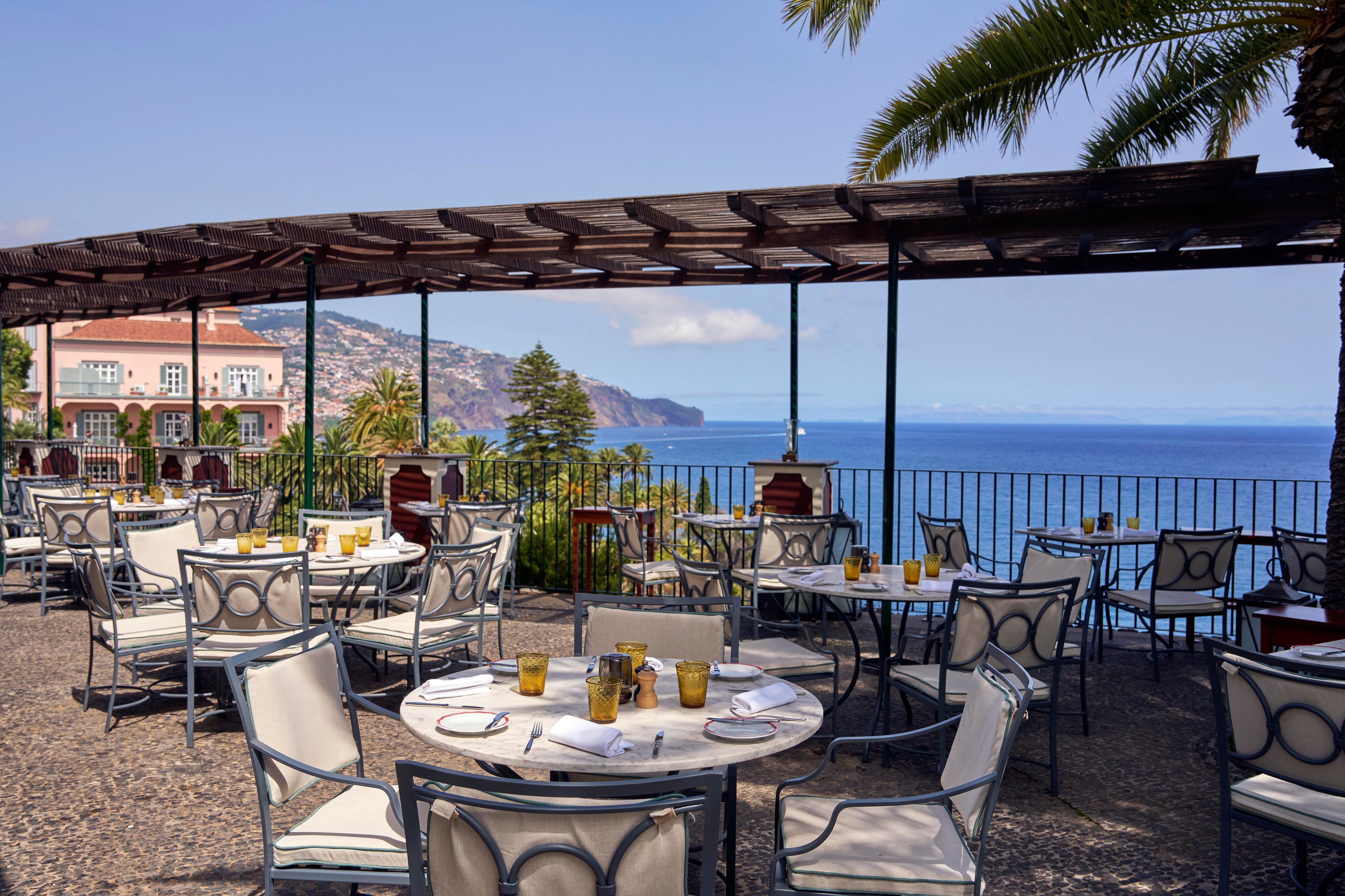 An outdoor restaurant terrace overlooks a calm blue ocean and distant coastline. White cushioned chairs surround round tables set with glasses and tableware under a wooden pergola. Palm trees and lush greenery frame the scene, while wrought-iron railings border the edge of the terrace. The atmosphere is bright and relaxed, suggesting a warm, sunny day in a coastal destination.