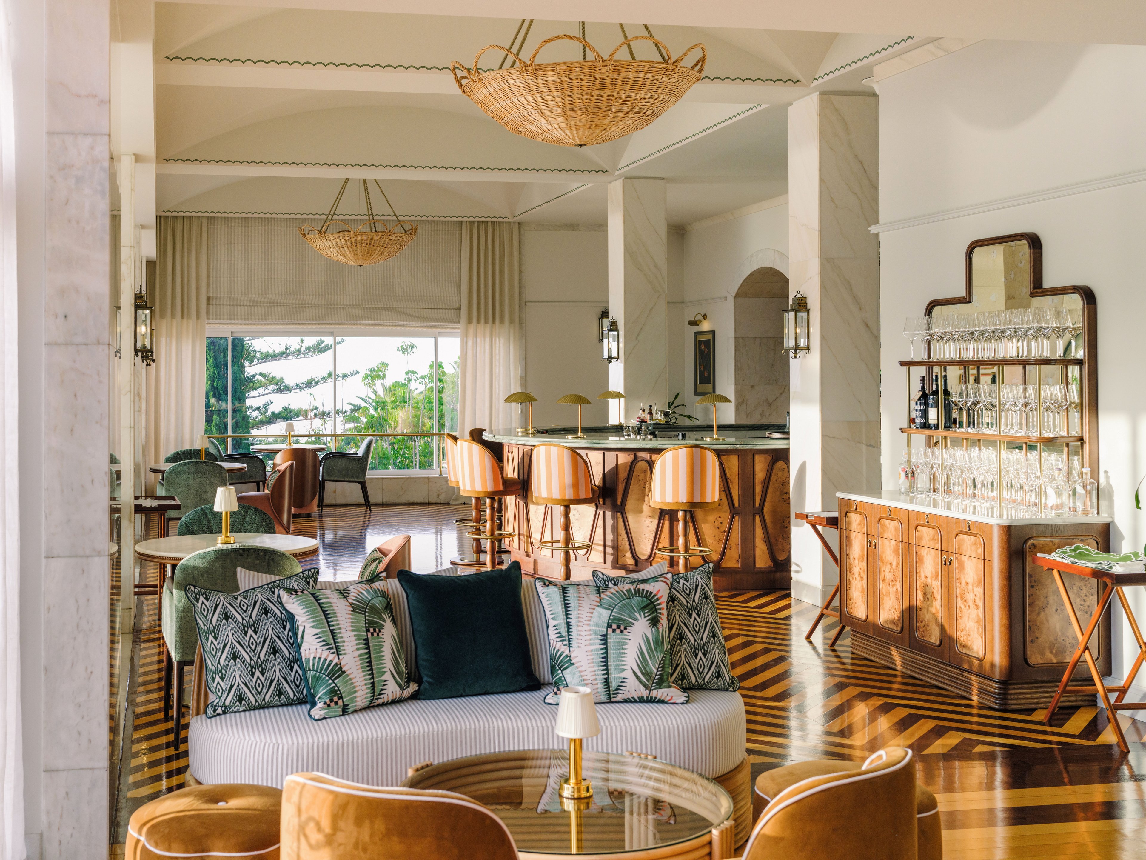 A spacious, sunlit hotel lounge features a stylish bar area with high stools and a mix of modern and vintage furniture. Plush armchairs and a patterned sofa are arranged around low tables, creating an inviting seating area. Large windows at the back reveal greenery and trees outside, enhancing the airy atmosphere. Warm wood floors, neutral walls, and woven light fixtures contribute to a sophisticated yet relaxed design.