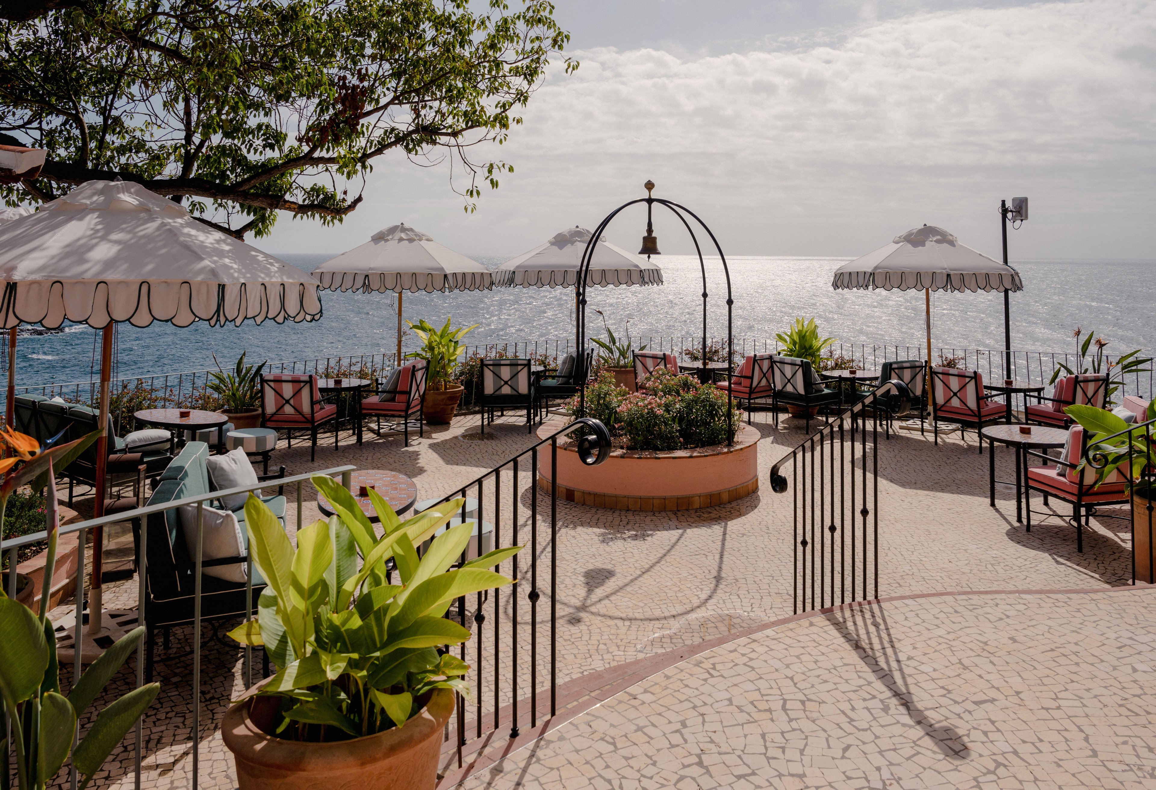 An open-air terrace café overlooks a calm ocean under a partly cloudy sky. White parasols shade red cushioned chairs and tables arranged along a curved railing. Potted plants and greenery frame the seating area, adding a tropical touch to the stone-paved patio. The scene feels relaxed and inviting, ideal for leisurely outdoor dining by the sea.