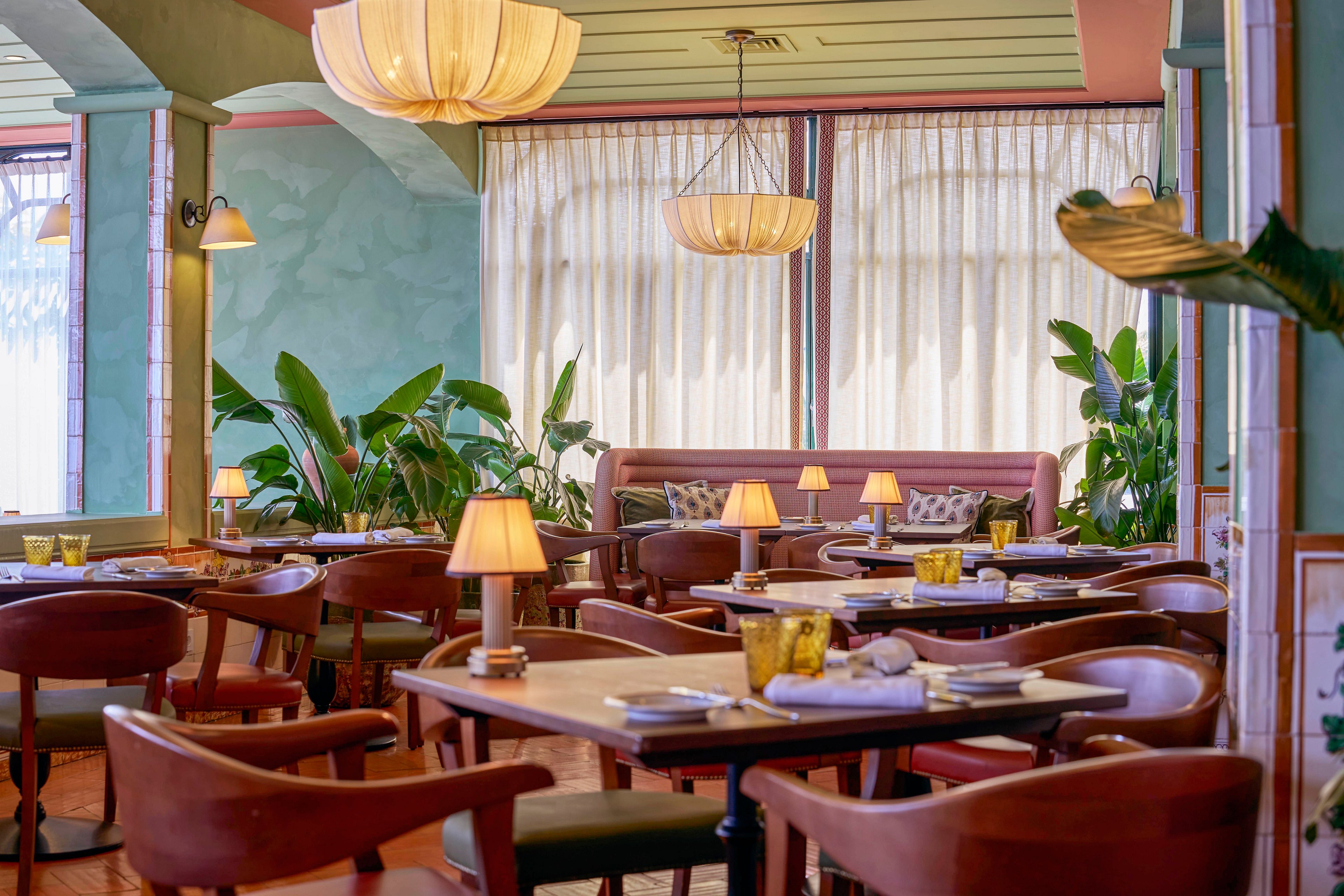 A stylish restaurant dining room features neatly arranged wooden tables and chairs. Soft pastel walls, large windows with sheer curtains, and lush green plants create a welcoming atmosphere. Warm table lamps and pendant lights cast a gentle glow over the space. The overall setting feels calm, inviting, and ready for guests.