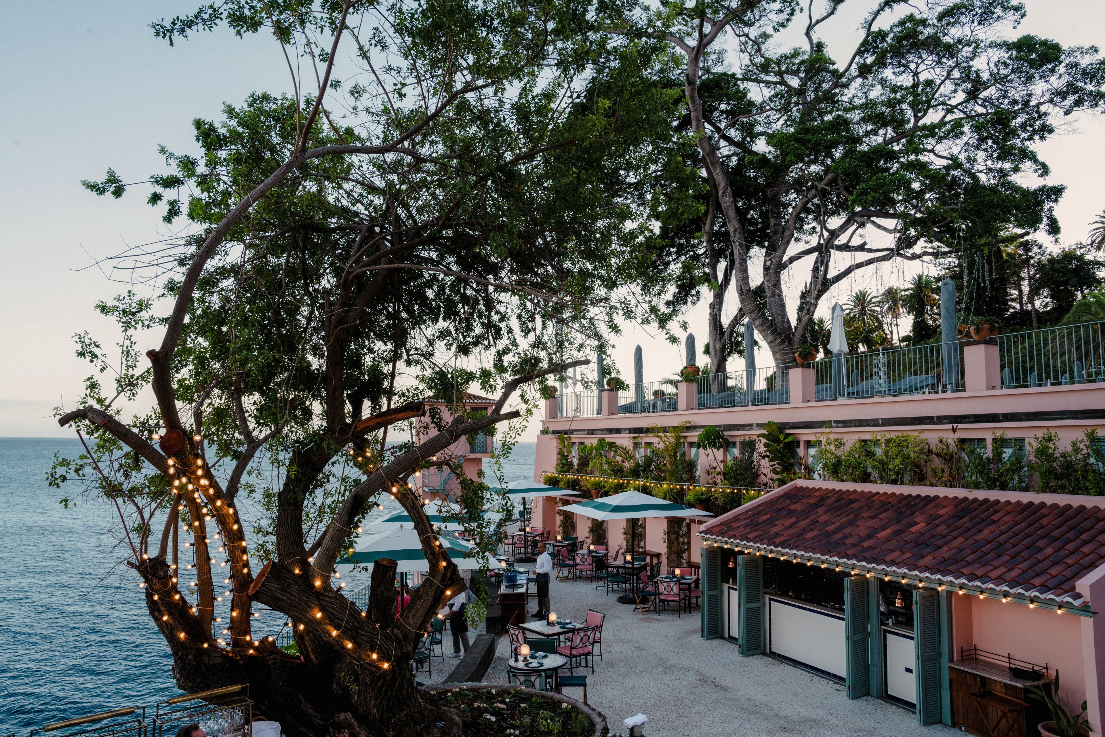 An open-air restaurant terrace overlooks a calm seaside at dusk. Diners sit under umbrellas and string lights wrapped around a large tree, creating a cozy atmosphere. The multi-level building features pastel walls, tiled roofs, and railings lined with greenery. The scene captures a relaxed coastal evening with soft natural light and gentle ocean views.