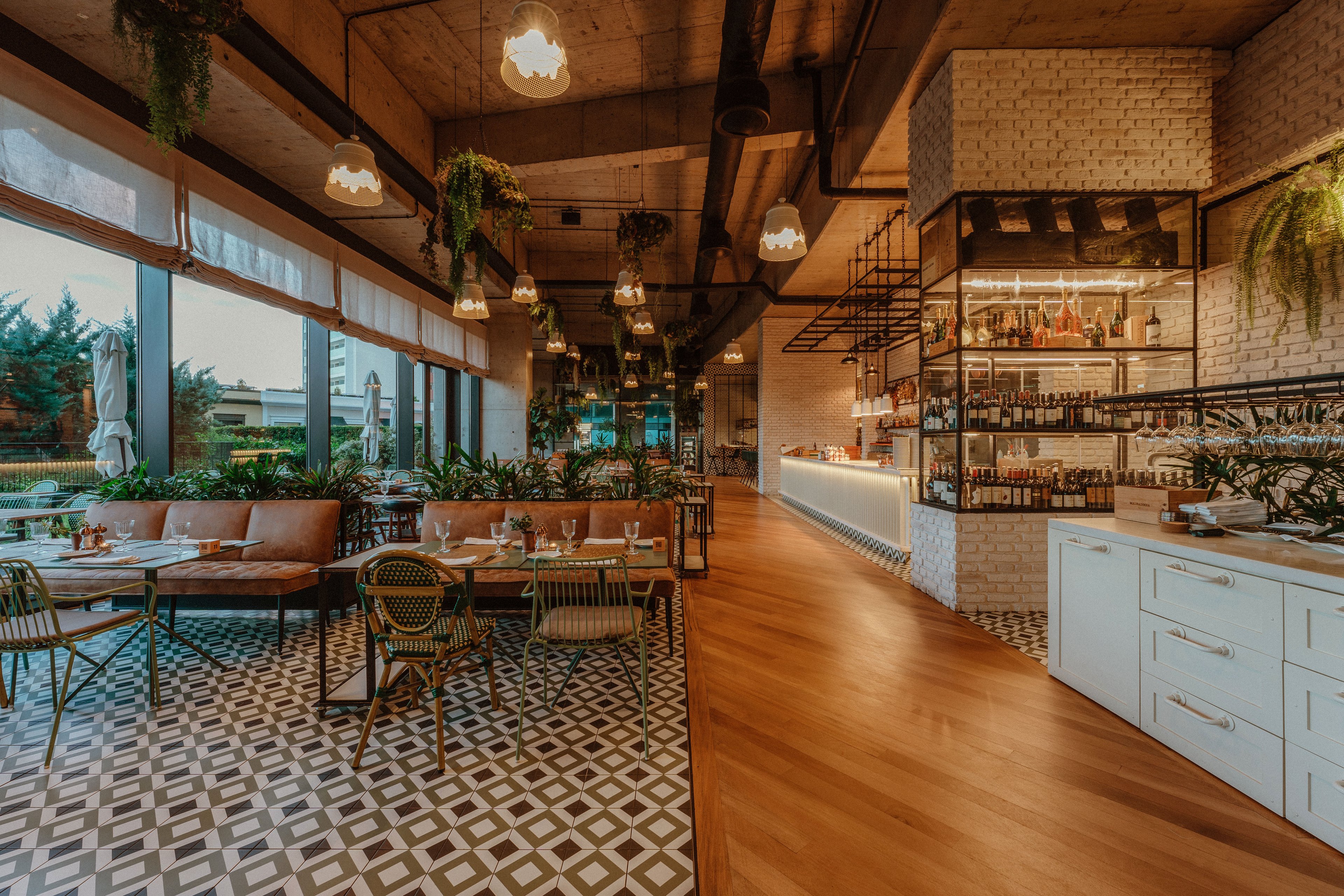Contemporary restaurant interior featuring a mix of wooden flooring and geometric patterned tiles. Large windows line one side, offering views of greenery and outdoor seating. A long bar with illuminated shelving displays bottles and glassware, while tables are set with glasses and cutlery. Hanging plants, pendant lights, and neutral tones create a cozy, inviting atmosphere.