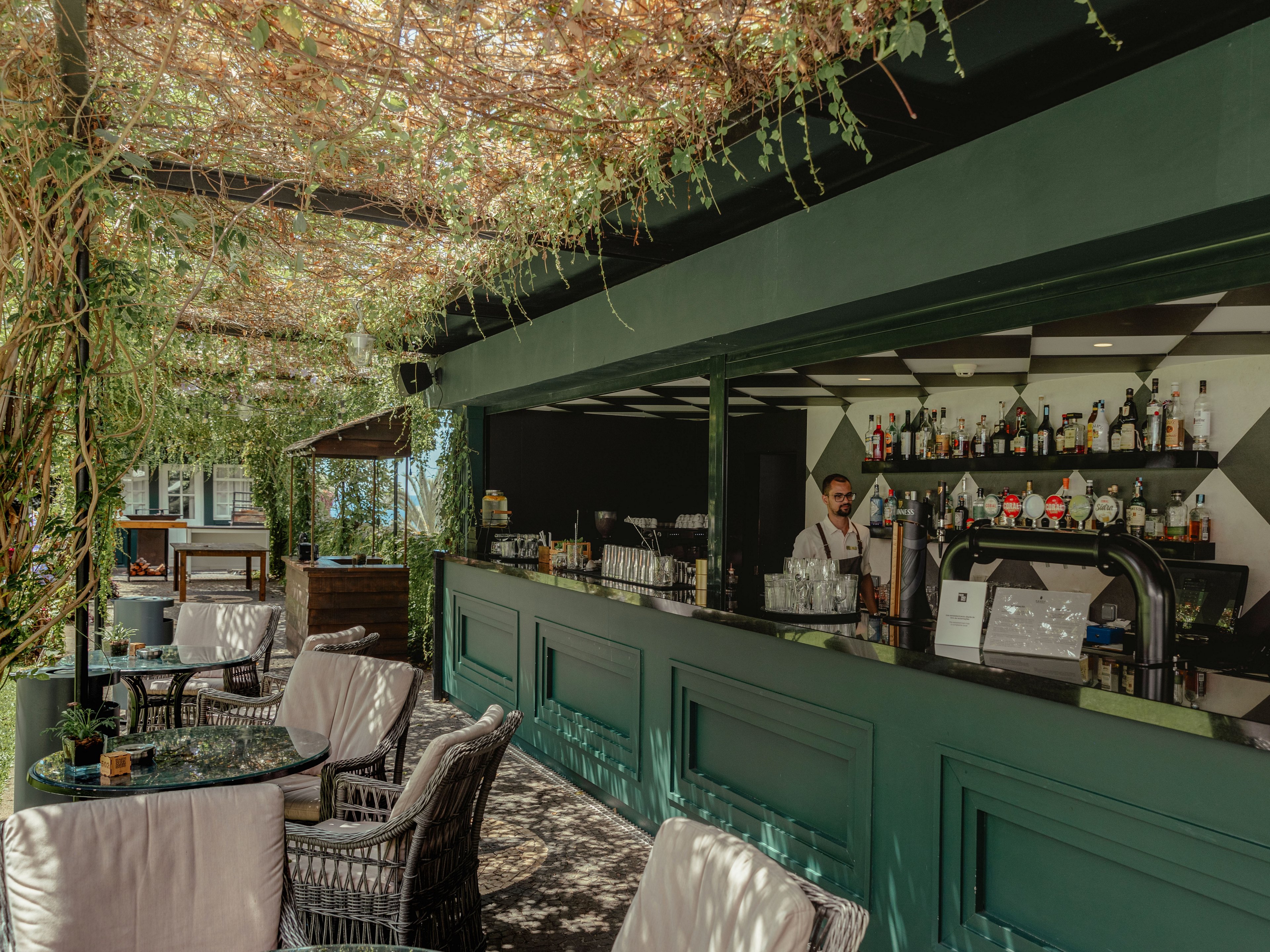 An outdoor bar area features a bartender standing behind a long green counter lined with bottles and glassware. Wicker chairs with light cushions surround glass-top tables in the foreground, creating a comfortable seating space. Overhead, dense greenery and vines form a natural canopy that filters warm sunlight across the scene. The overall atmosphere is relaxed and inviting, suitable for socializing and enjoying drinks in a garden-like setting.