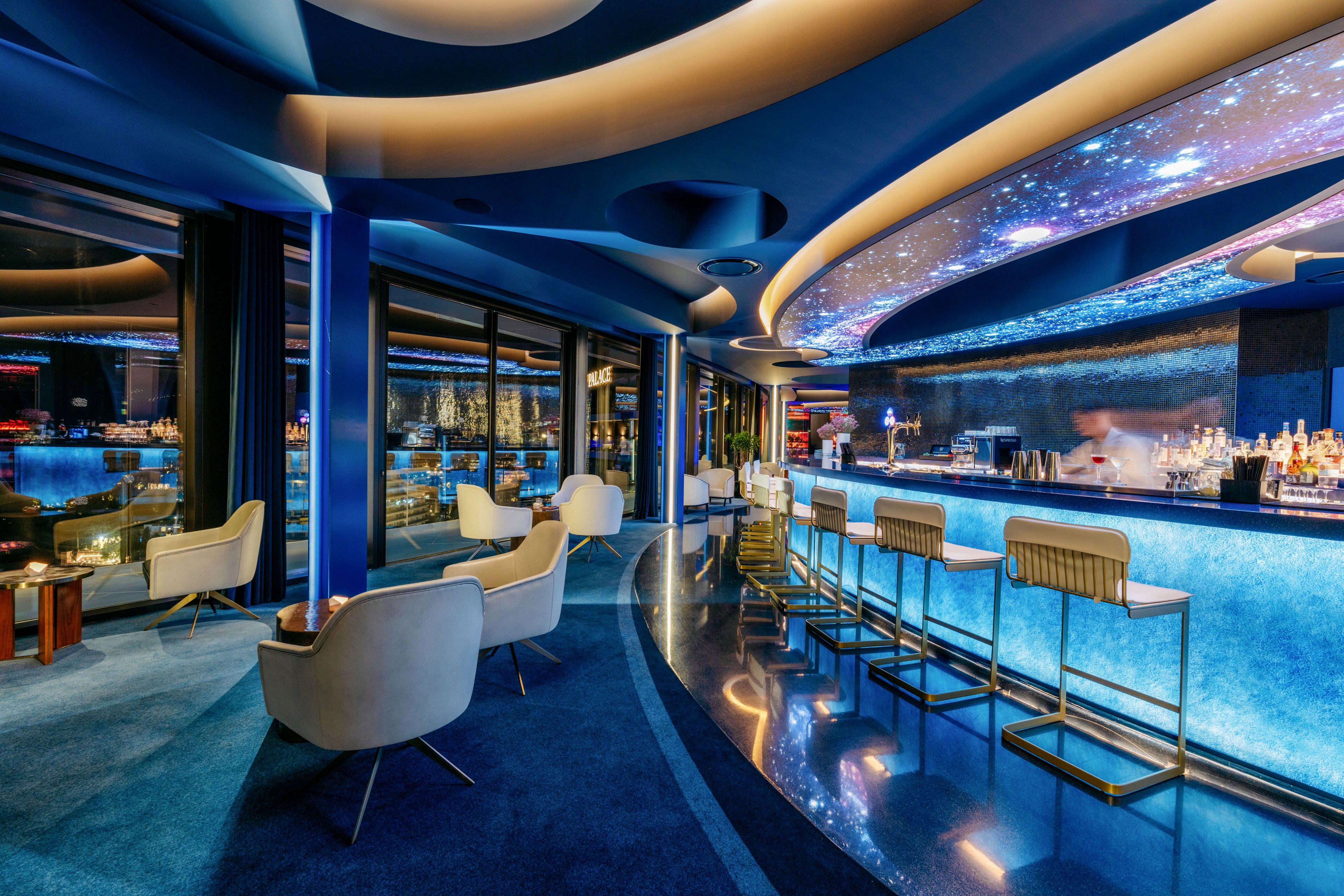 A modern lounge bar interior features sleek blue lighting and curved architectural lines. High stools line an illuminated bar counter, with bottles and glassware arranged behind it. Comfortable white armchairs and low tables sit near large windows overlooking a nighttime city skyline. The ceiling showcases a cosmic, starry design that enhances the futuristic atmosphere.