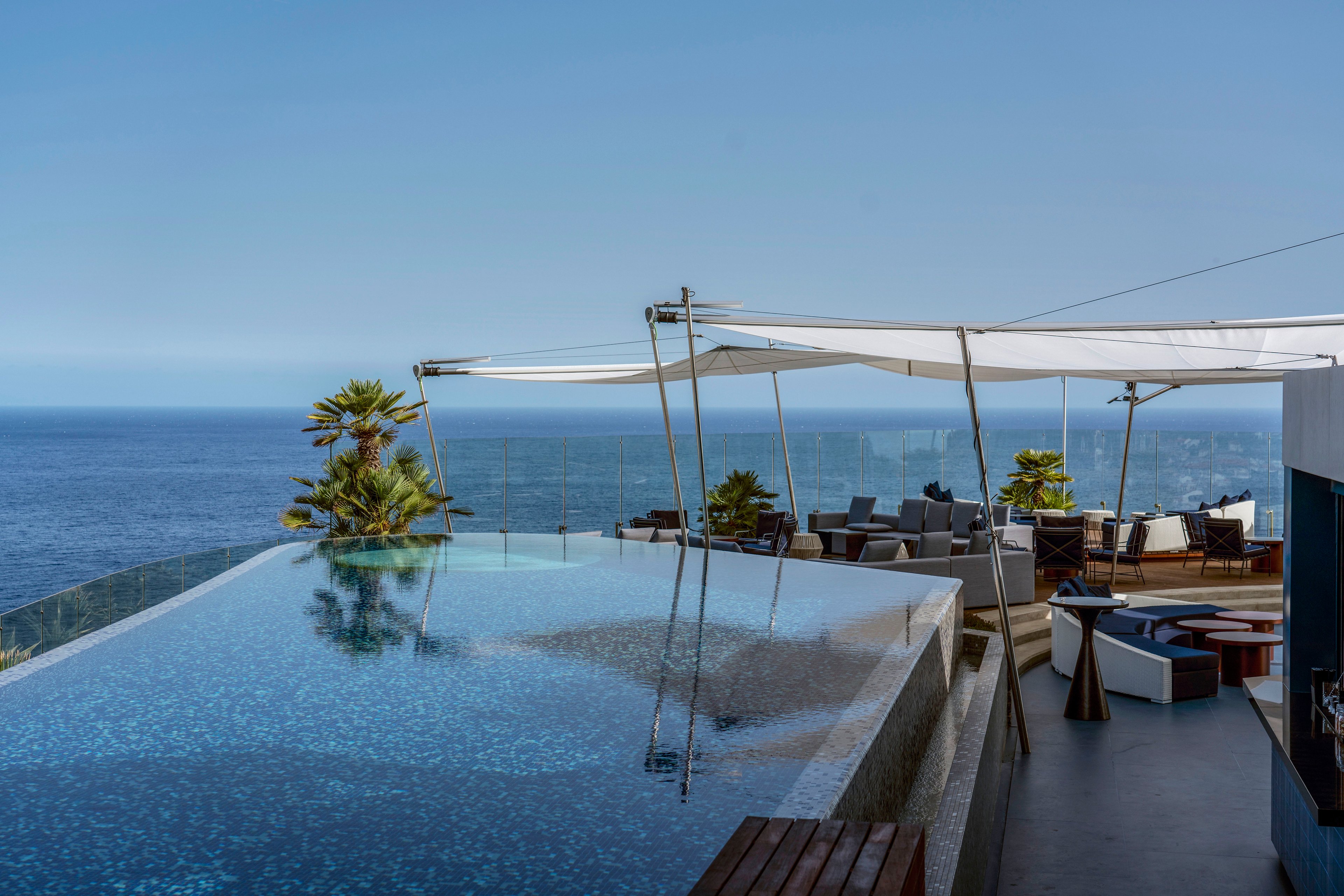 A modern infinity pool stretches toward a vast blue ocean horizon, creating a seamless connection between water and sea. The terrace features contemporary outdoor seating under white shade sails, suggesting a high-end resort or private villa setting. Glass railings frame the view while palm plants add a touch of greenery to the sleek architectural design. The clear sky and calm water convey a tranquil, relaxing atmosphere ideal for leisure and vacation.