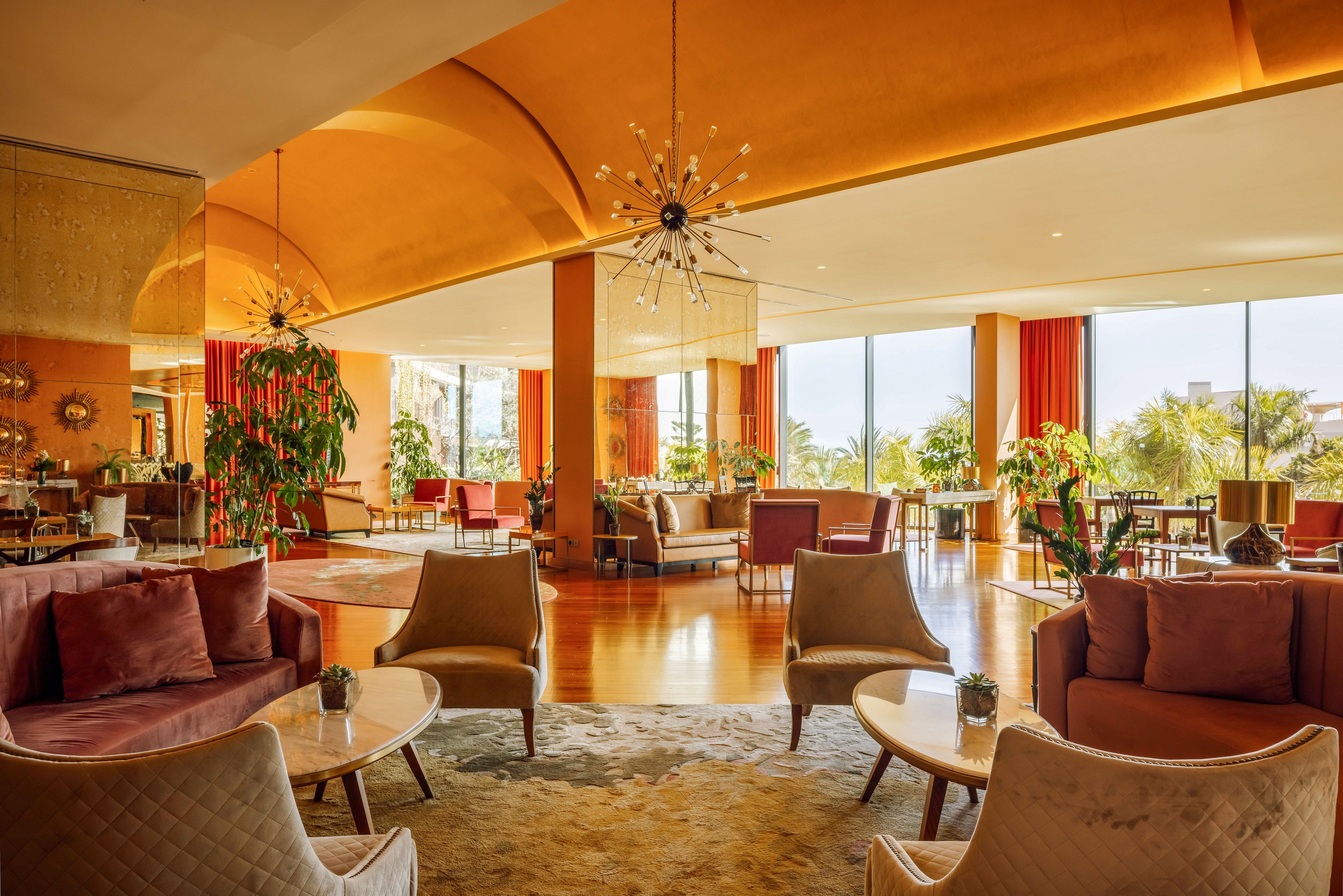 Spacious hotel lounge with contemporary armchairs and sofas arranged around low coffee tables. Large floor-to-ceiling windows fill the room with natural light and reveal lush greenery outside. Warm orange and beige tones dominate the interior, complemented by red curtains and wooden flooring. Decorative plants, modern chandeliers, and small table accents create an inviting, upscale atmosphere.