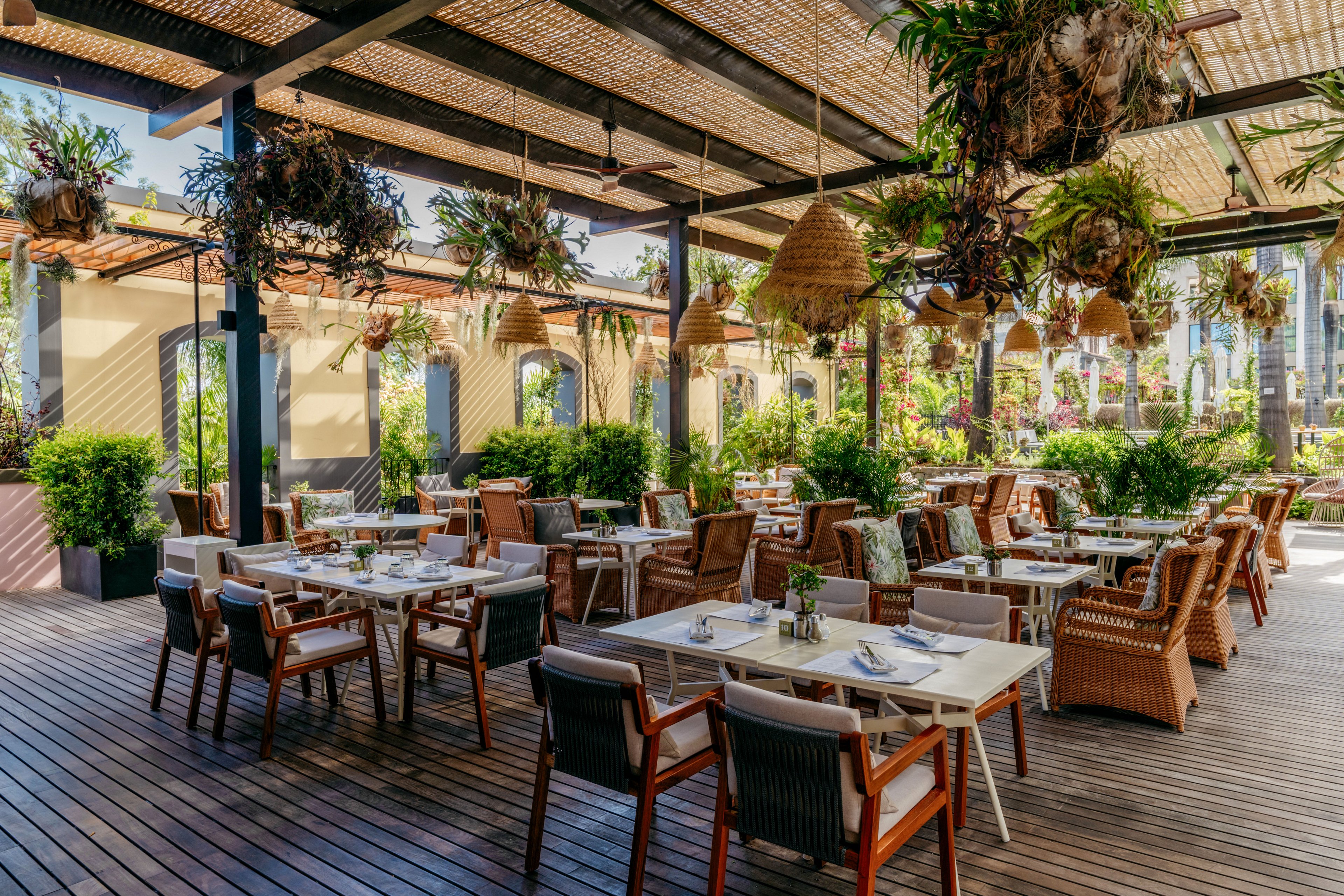 An inviting outdoor restaurant terrace features neatly arranged tables and wooden chairs under a woven canopy. Hanging plants and wicker lamps add natural texture and warmth to the space. Lush greenery surrounds the seating area, creating a relaxed garden atmosphere. The setting appears ready for guests, with tableware and glassware already placed.