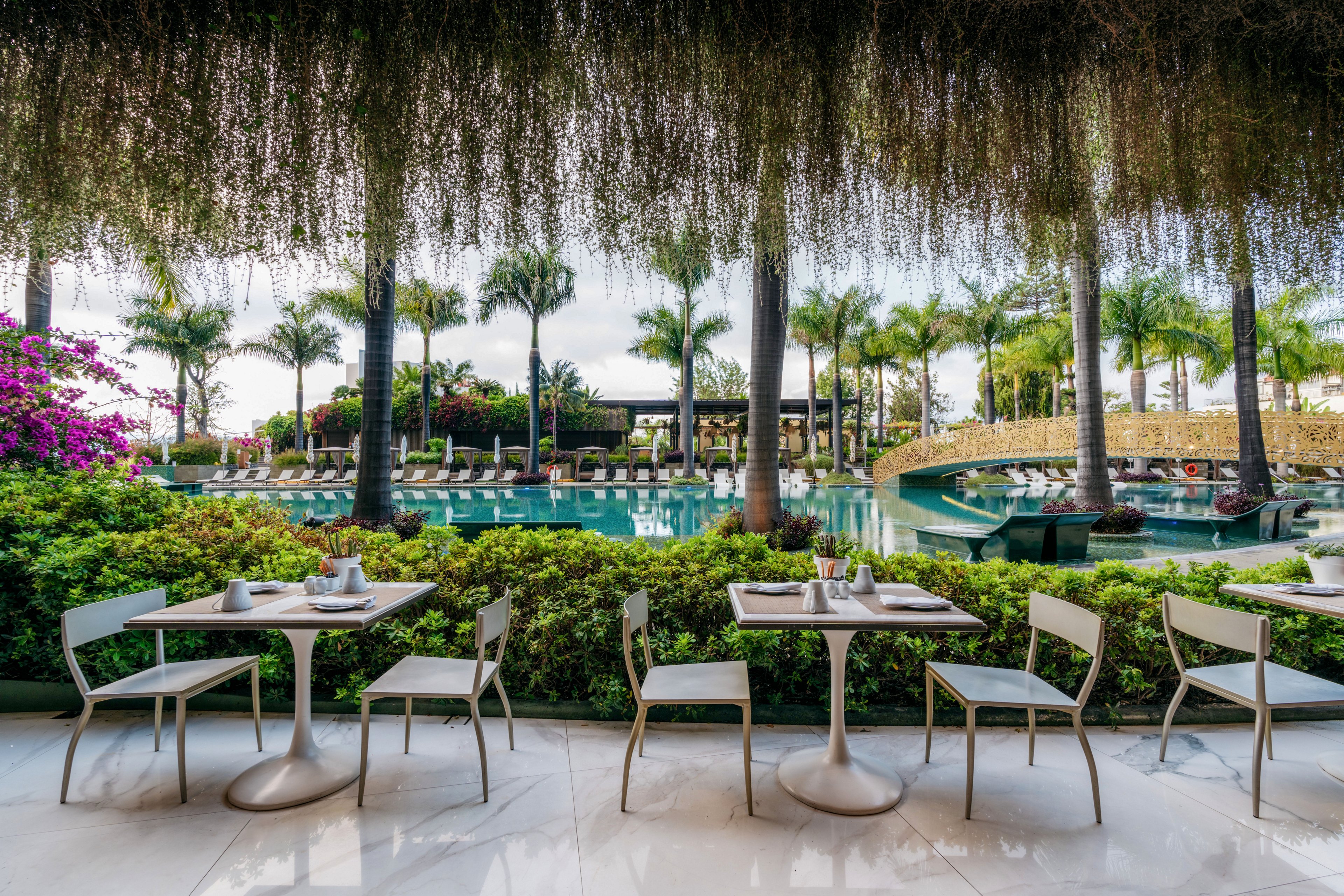 An open-air café terrace with modern white tables and chairs faces a large resort swimming pool. Lush green hedges, hanging plants, and palm trees frame the view, creating a tropical atmosphere. Sun loungers and umbrellas line the poolside area in the background, suggesting a relaxing vacation setting. The scene feels calm and inviting, ideal for outdoor dining and leisure.
