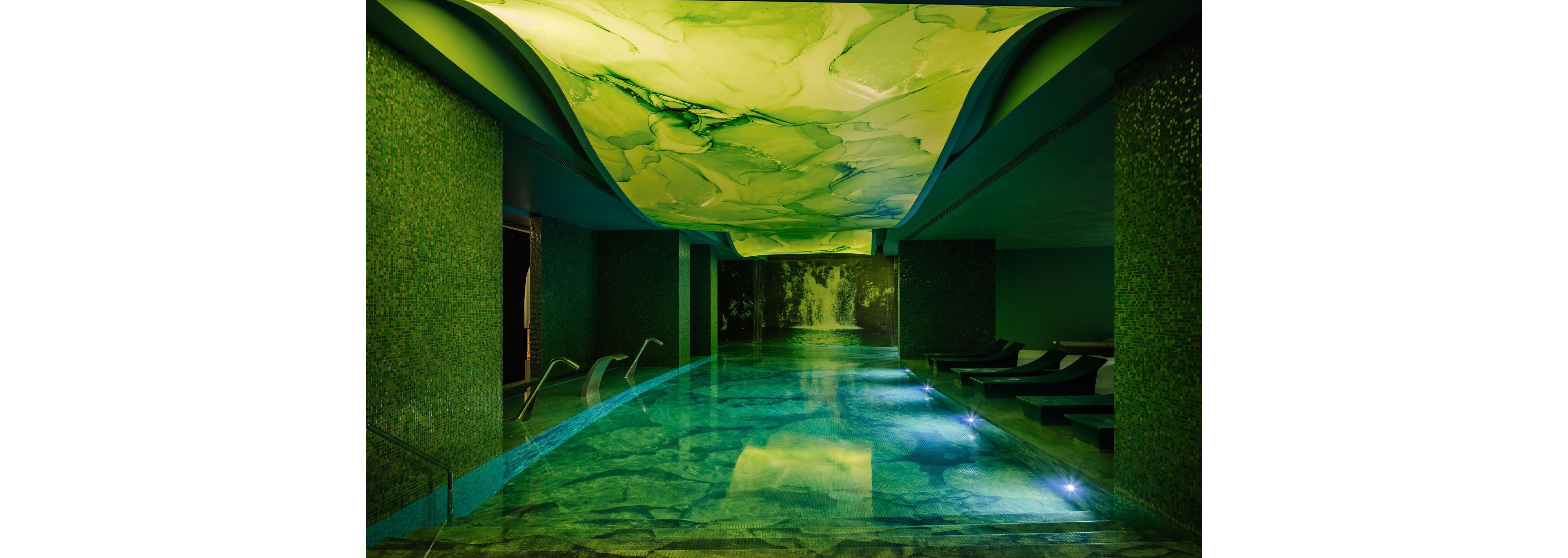An indoor spa pool glows with dramatic green lighting and a marbled ceiling design. Lounge chairs line the right side of the pool, creating a relaxing retreat atmosphere. Soft underwater lights illuminate the water, leading toward a wall mural of a waterfall in the background. The overall scene feels tranquil, modern, and immersive.