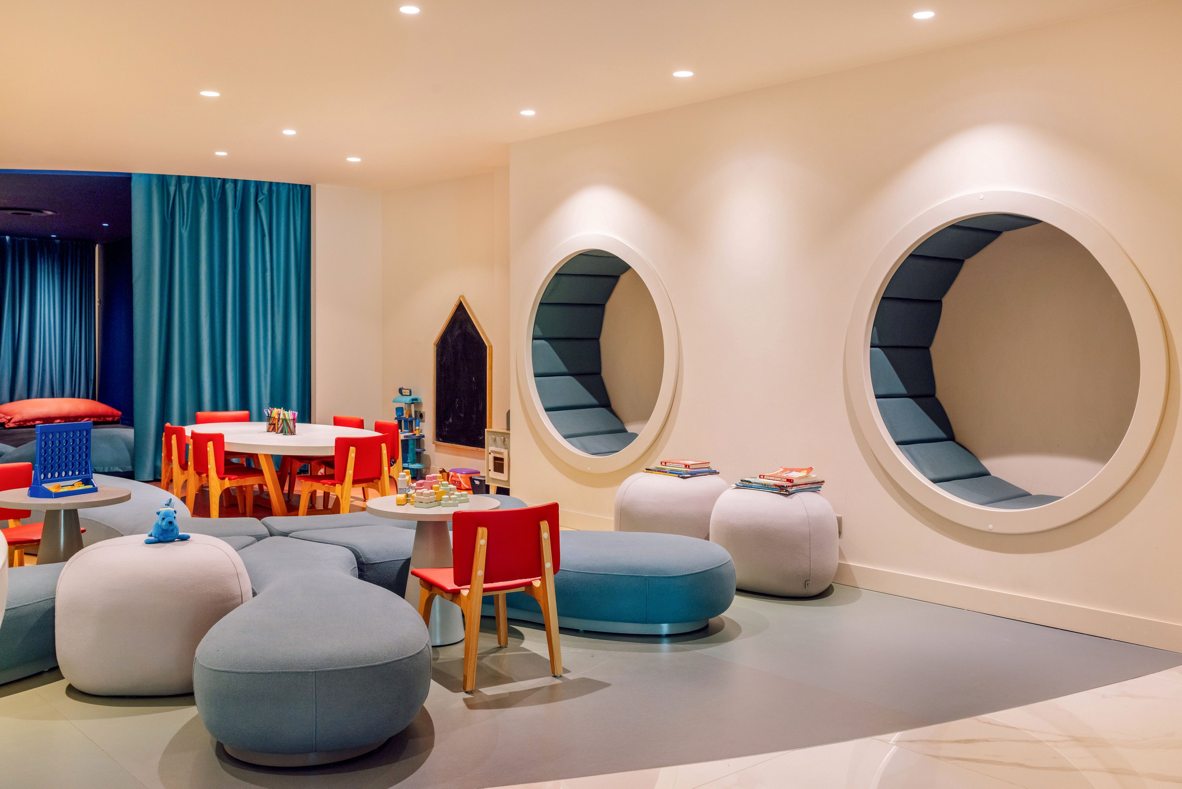 A contemporary children’s playroom features round cushioned seating and built-in circular wall nooks. Bright red and orange chairs surround low tables with toys and books scattered on top. Soft blue poufs and curved benches create a cozy, informal lounge area. Turquoise curtains and warm recessed ceiling lights add a playful yet relaxing atmosphere.
