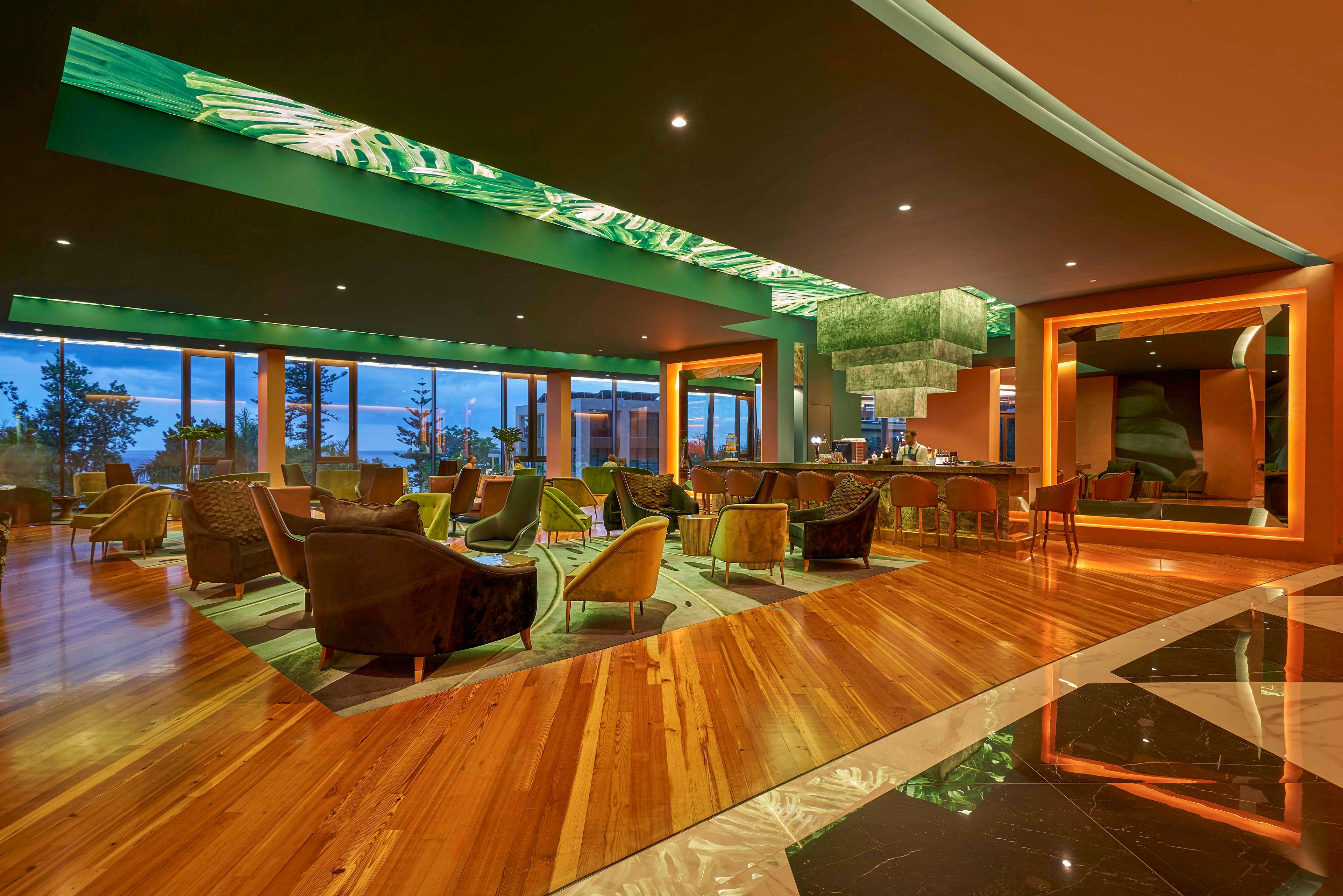 A contemporary hotel lounge features plush armchairs and low tables arranged across a polished wooden floor. Large windows reveal an evening view of the outdoors, adding depth and natural light to the space. The ceiling and bar area are accented with green illuminated panels and geometric design elements. Warm orange and gold tones create a cozy yet upscale atmosphere suitable for socializing or relaxing.