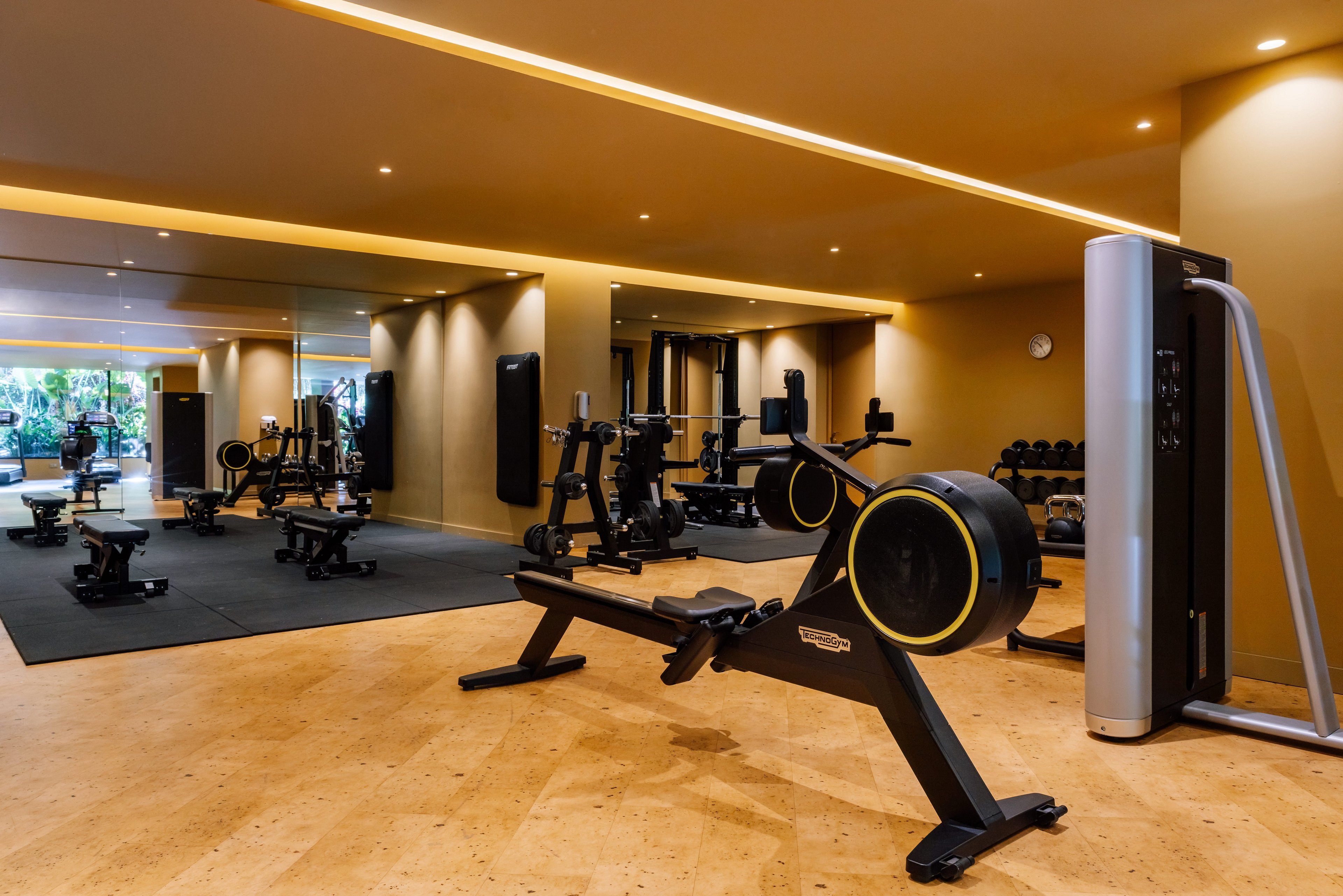 A contemporary fitness center features a rowing machine in the foreground and various strength-training equipment in the background. The room has warm lighting, wooden flooring, and large mirrors that visually expand the space. Free weights, benches, and resistance machines are arranged neatly on a dark mat area. Natural light enters from glass doors at the far end, creating an inviting atmosphere for workouts.