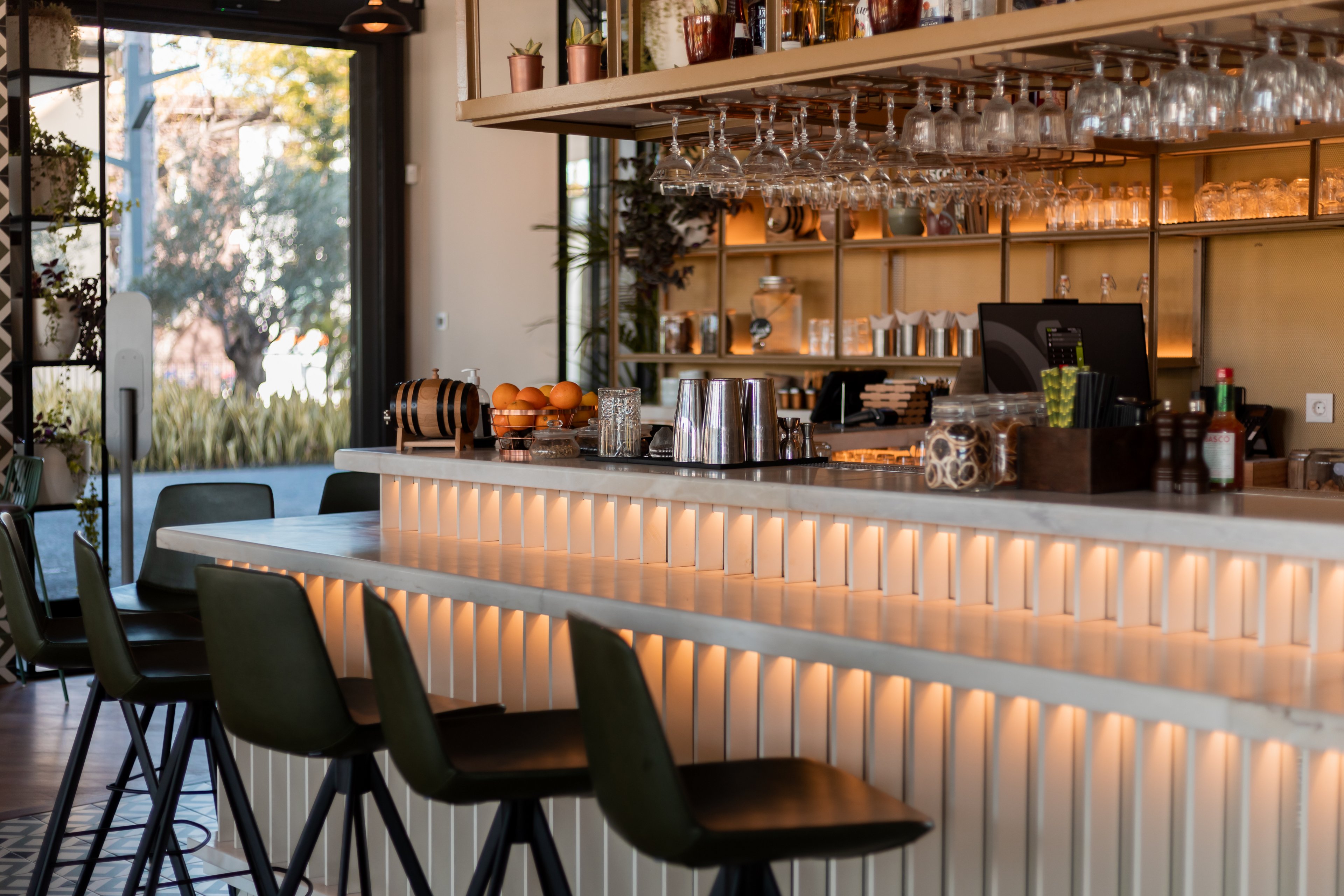 Contemporary cafe bar interior with a sleek counter and high green bar stools. Warm under-counter lighting and hanging glassware create an inviting atmosphere. Shelves in the background display bottles, plants, and decor items. Large windows let in natural daylight and offer a view of greenery outside, enhancing the relaxed, modern setting.
