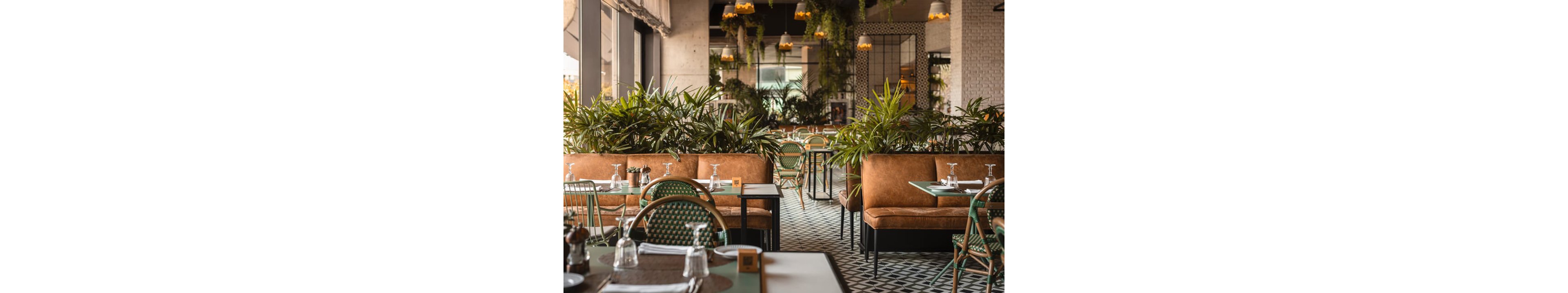 A contemporary restaurant dining area features tan upholstered booths and green woven chairs separated by tall indoor plants. Tables are neatly set with upside-down wine glasses, cutlery, and placemats, ready for guests. Large windows on the left bring in natural light, while hanging lamps and ceiling greenery add warmth and texture. The patterned floor tiles and mix of materials create a stylish, inviting atmosphere.