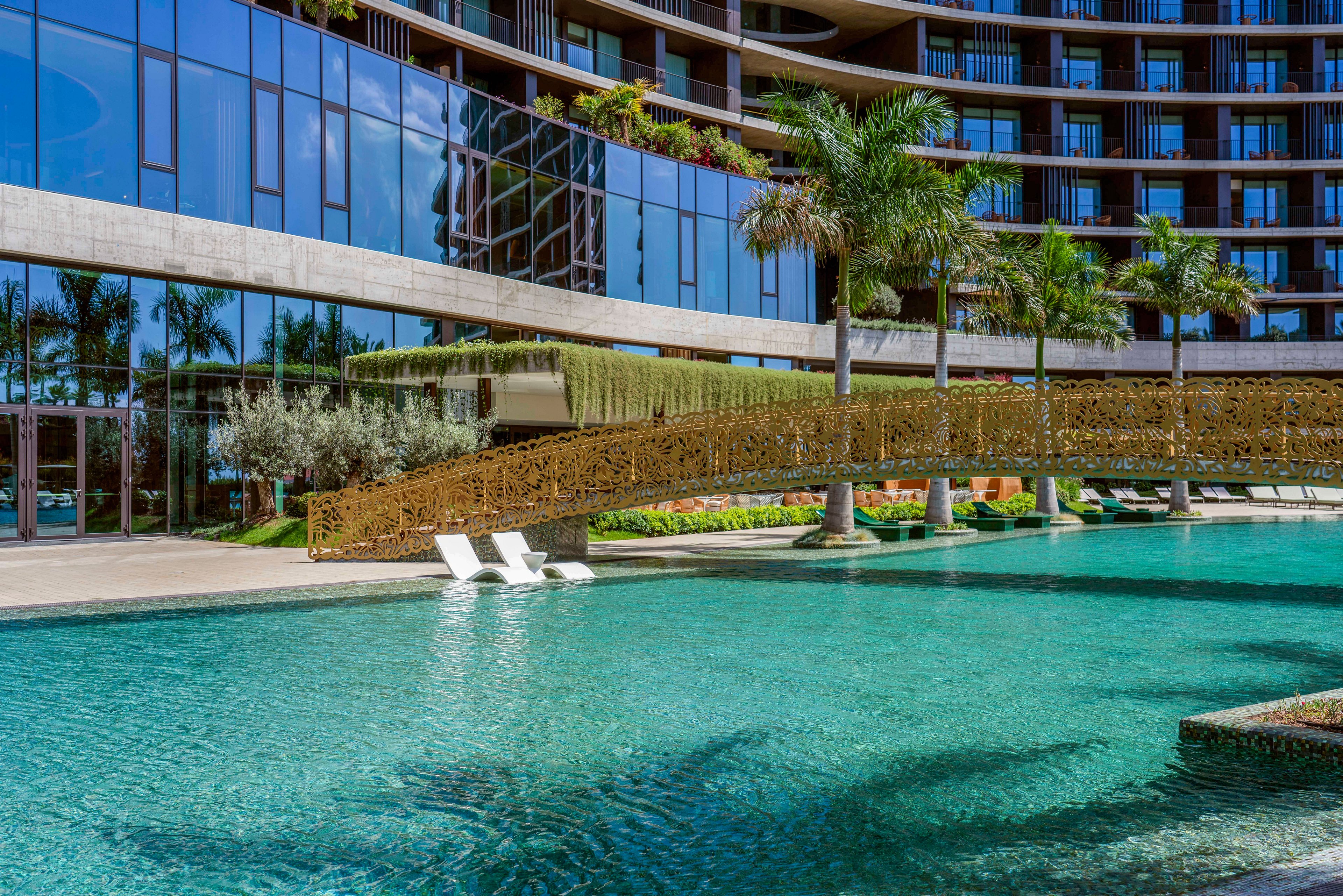 A contemporary resort features a large turquoise swimming pool in front of a curved glass-fronted building. A decorative pedestrian bridge spans the pool, with tropical palm trees and greenery integrated into the design. Two white lounge chairs sit partially submerged in the shallow water, inviting relaxation. Reflections of the architecture and plants shimmer on the pool surface, creating a luxurious vacation atmosphere.