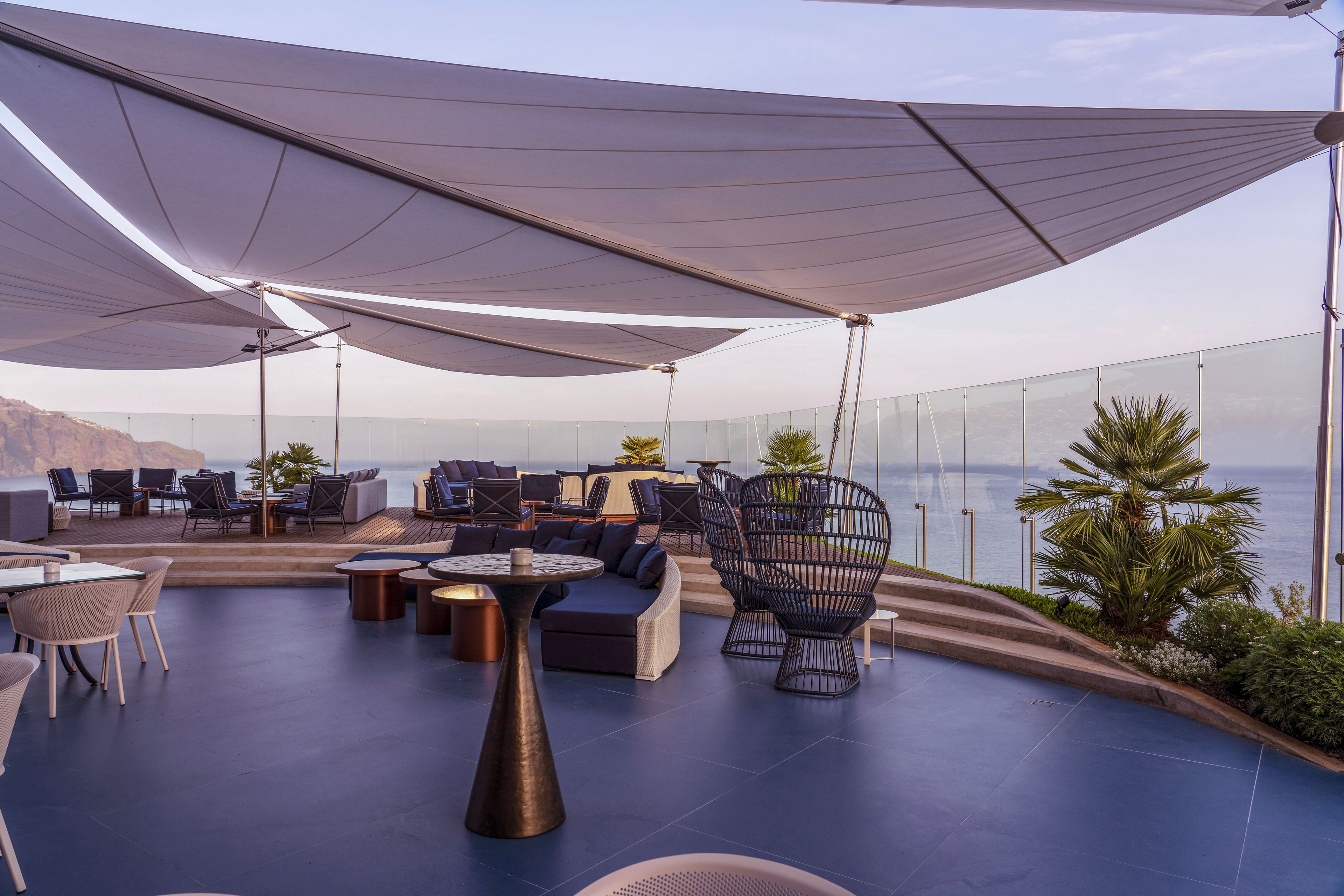 Contemporary outdoor lounge seating is arranged on a rooftop terrace with expansive ocean views. Large sail-like shade canopies stretch overhead, creating a stylish and comfortable environment. Glass railings line the edge of the terrace, and potted palm trees add greenery to the modern design. The soft evening light casts a warm, relaxed atmosphere across the space.