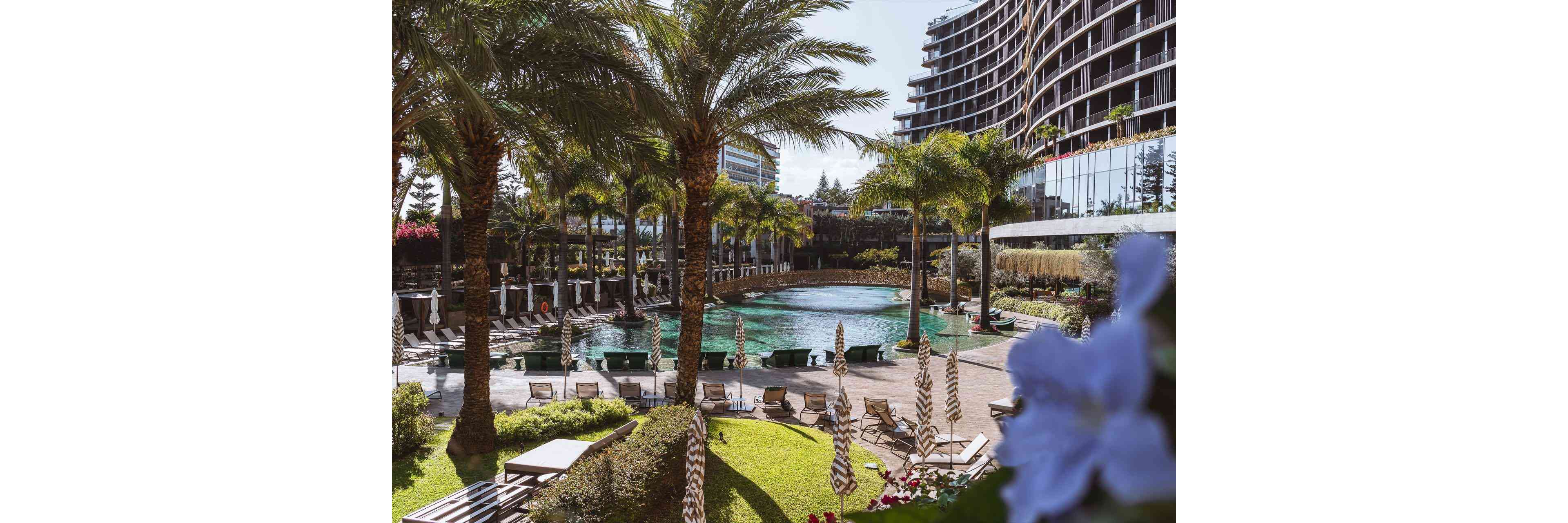 A large resort swimming pool is framed by tall palm trees and sun loungers in a landscaped courtyard. Curved modern hotel buildings rise in the background, reflecting the sunny outdoor setting. Parasols, manicured lawns, and flowering plants add to the relaxed vacation atmosphere. The scene suggests a warm, tropical destination ideal for leisure and relaxation.