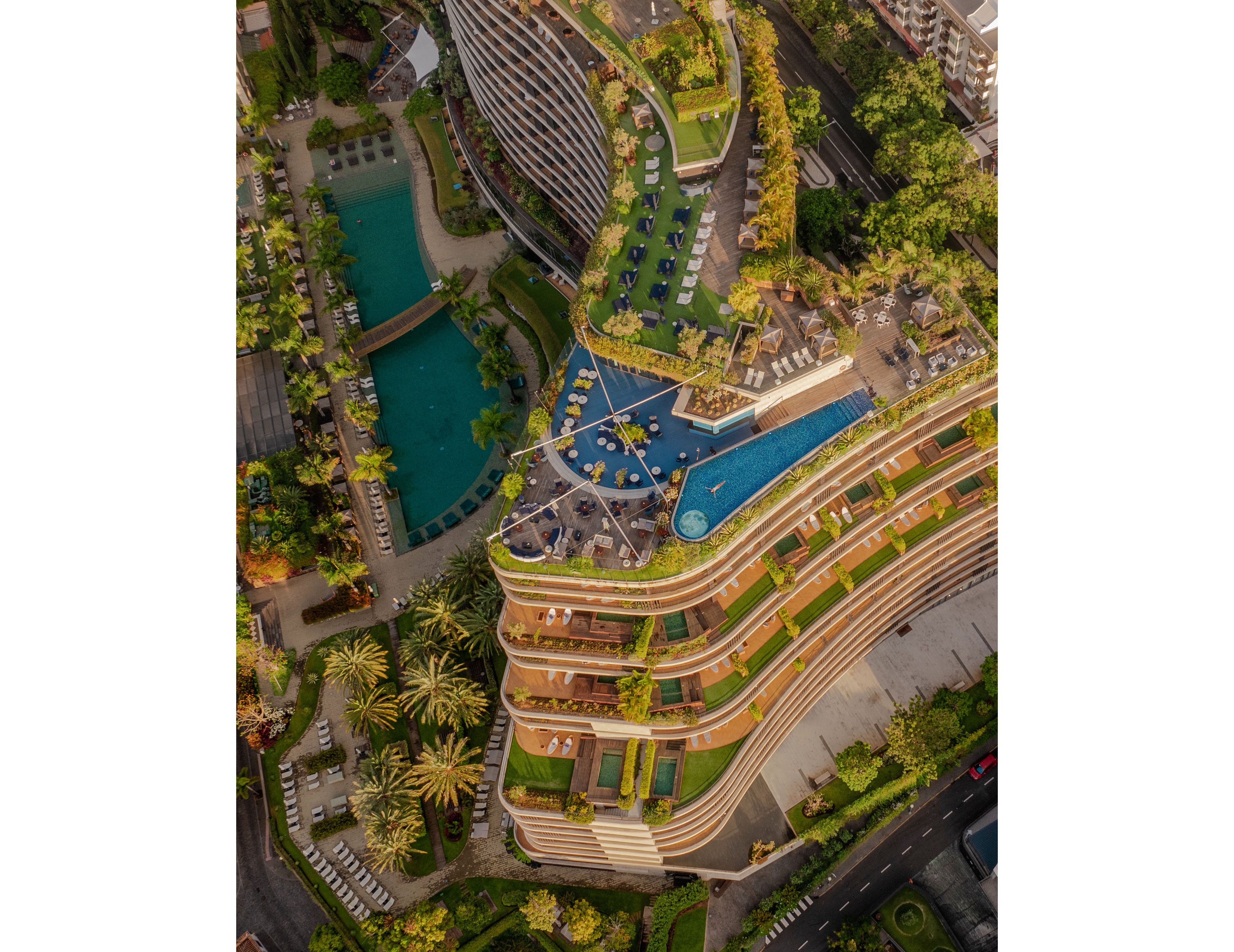 A contemporary multi-level resort complex is captured from above, showcasing curved terraced architecture. Several outdoor swimming pools and sun loungers are arranged across different levels, surrounded by lush greenery and palm trees. The scene includes landscaped gardens, pathways, and adjacent urban streets, highlighting the contrast between nature and city. Warm sunlight enhances the textures of the building and the vibrant colors of the vegetation and water.