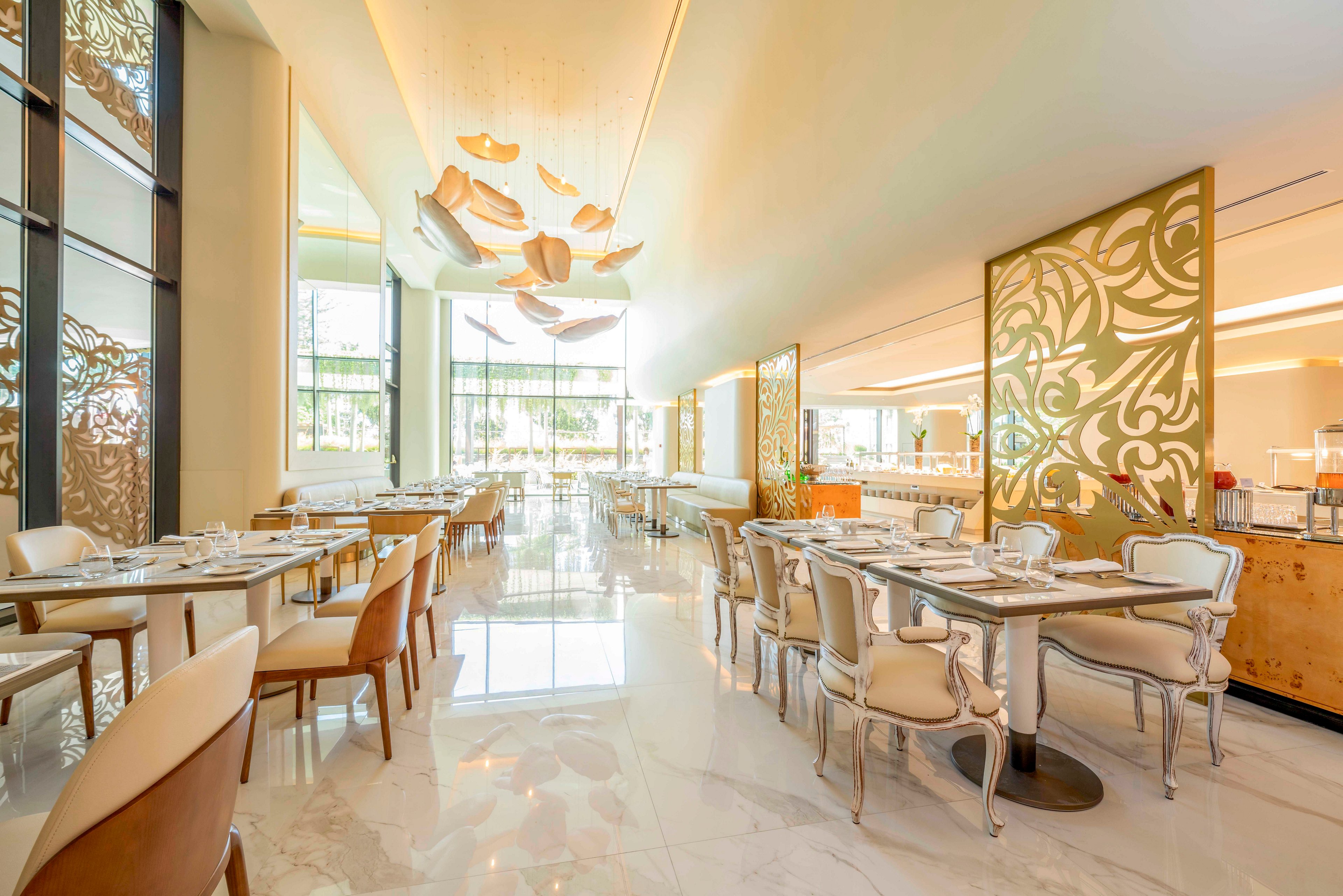 Spacious restaurant dining area with contemporary tables and upholstered chairs is shown in warm natural light. Large floor-to-ceiling windows open to an outdoor view, enhancing the airy atmosphere. Decorative ceiling fixtures and ornate partition panels add a touch of luxury and artistic detail. The polished marble floor and neatly set tables suggest a high-end, refined dining experience.