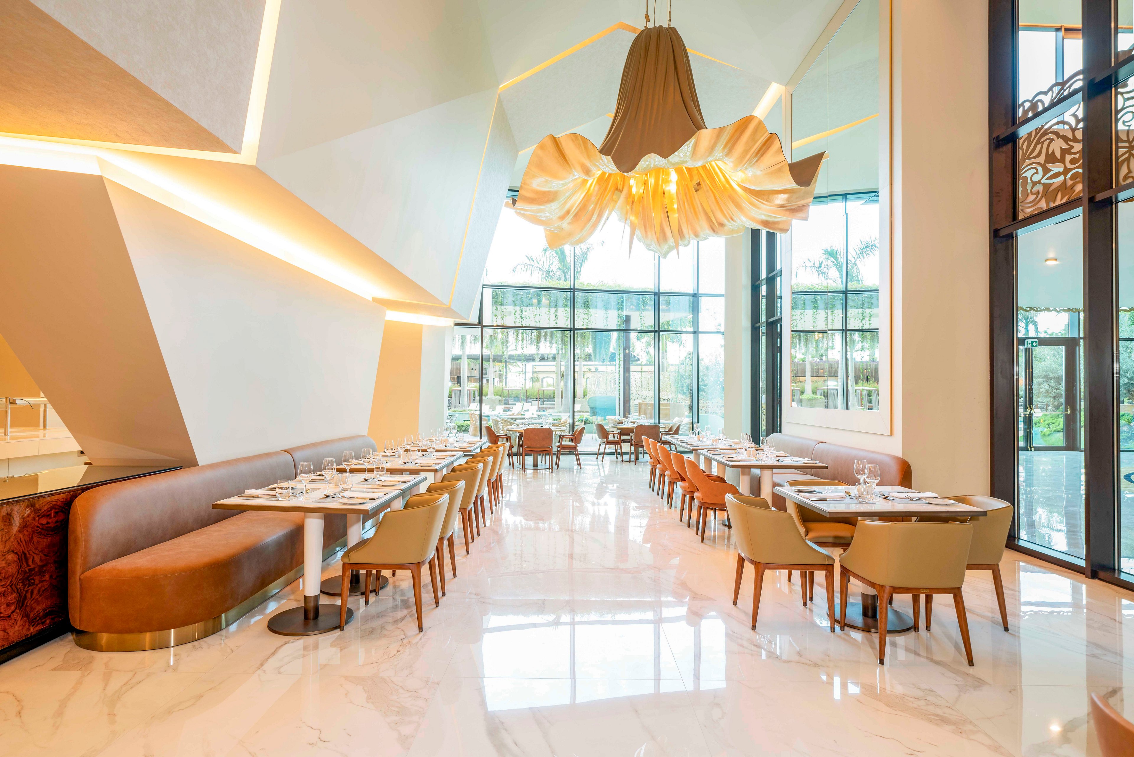 Upscale restaurant dining area featuring modern tables and upholstered chairs is shown in a bright, spacious interior. Large floor-to-ceiling windows reveal an outdoor view with palm trees and natural light flooding the space. A sculptural, flowing chandelier hangs from the high ceiling, complemented by geometric architectural details and warm accent lighting. Glossy marble-style flooring and neatly set tables create an elegant, inviting atmosphere.