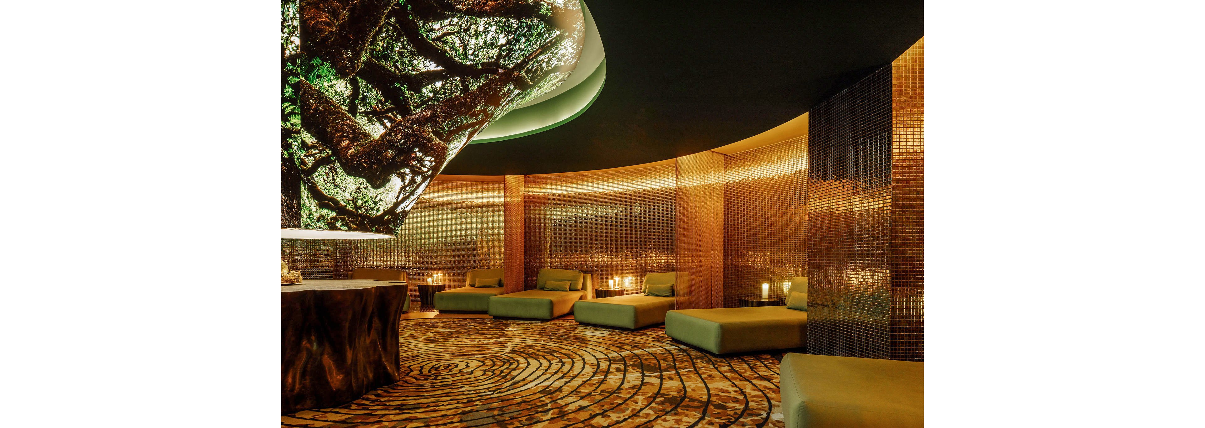 A luxurious spa lounge features multiple cushioned daybeds arranged around a circular room. Warm golden lighting reflects off textured metallic walls, creating a serene and intimate atmosphere. The ceiling showcases a large organic tree canopy image that enhances the connection to nature. Curved architectural lines and patterned flooring guide the eye through the tranquil relaxation space.