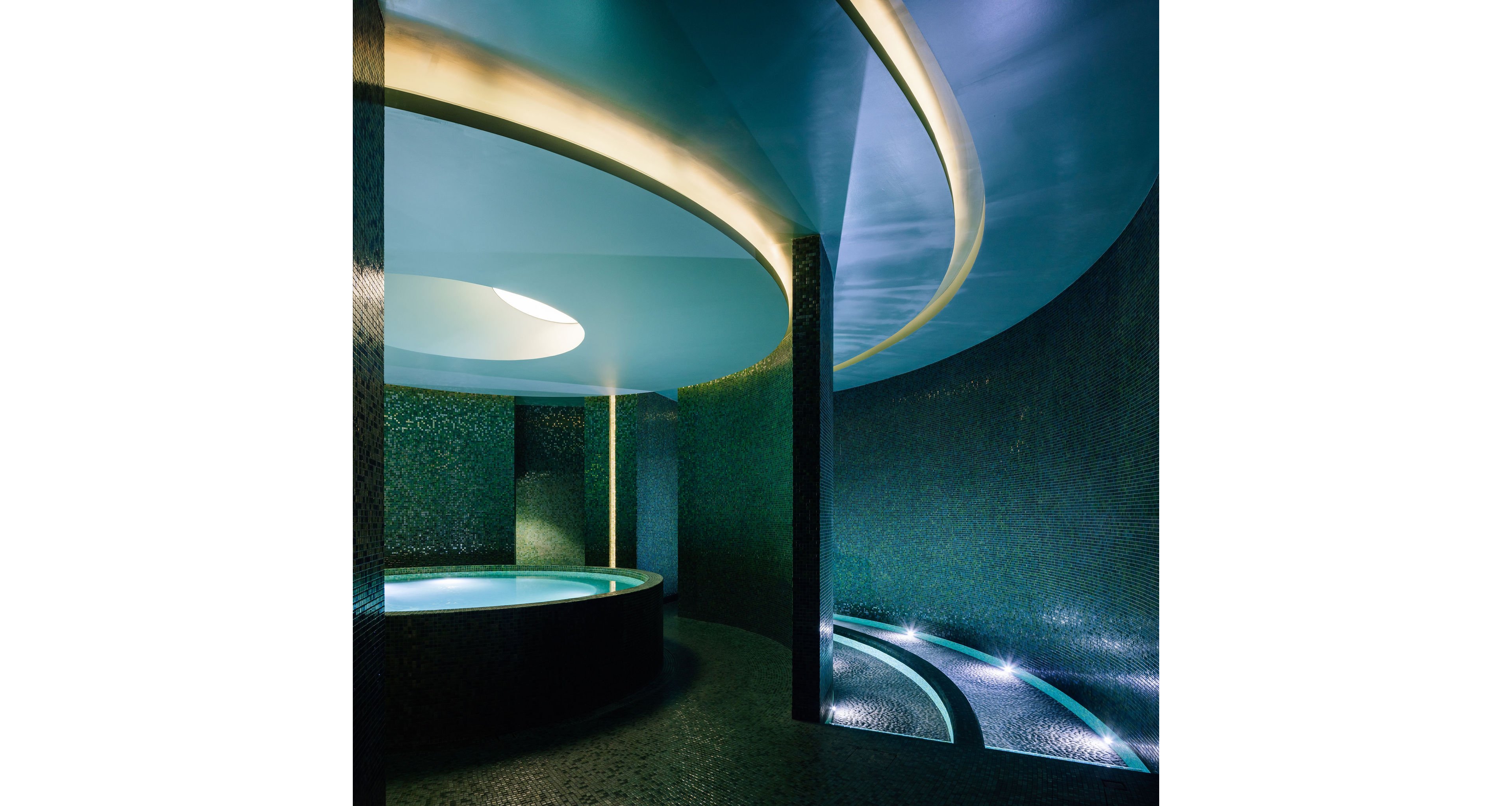 A circular indoor spa pool sits in a dramatic, modern architectural space. Curved walls and ceilings create a flowing tunnel-like environment around the water. Soft blue and green tones dominate the scene, enhanced by concealed strip lighting and reflections on mosaic tiles. The overall atmosphere is tranquil, luxurious, and slightly futuristic.