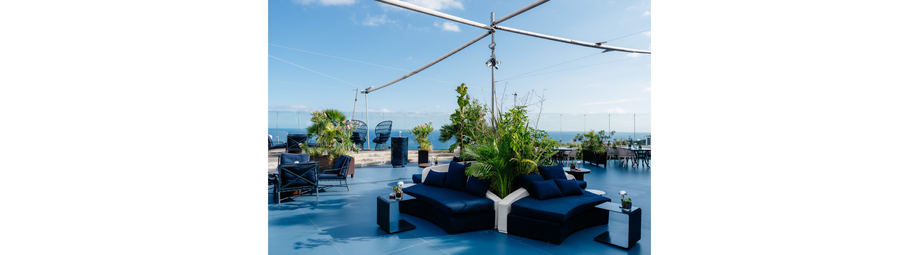 A contemporary rooftop terrace features low navy-blue lounge sofas arranged around lush green plants. Glass railings frame a clear view of the ocean under a bright blue sky with scattered clouds. Additional seating areas and potted greenery create a relaxed, open-air social space. The overall scene feels modern, airy, and inviting for outdoor gatherings.