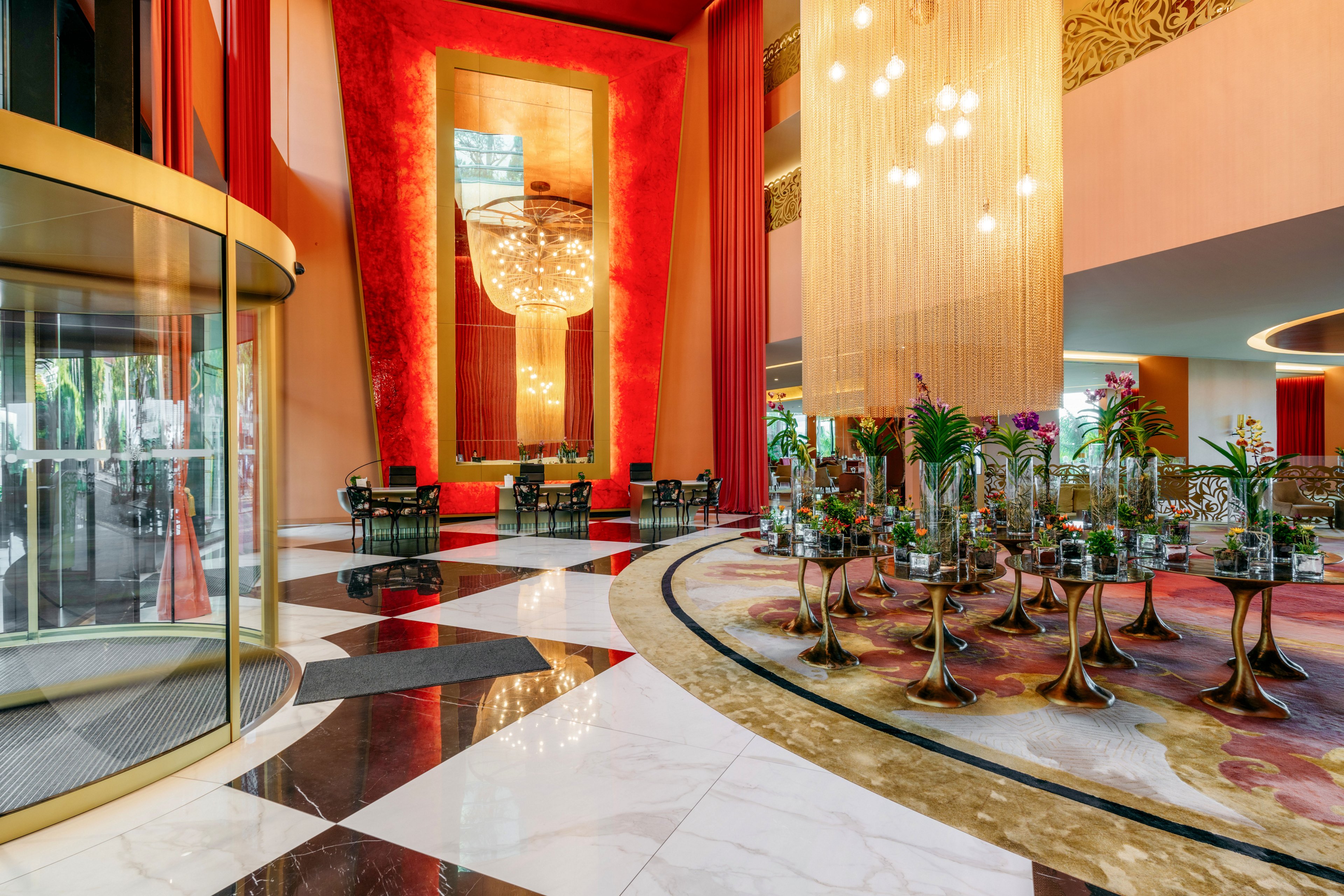 A spacious, upscale hotel lobby features a dramatic red accent wall and a large mirrored panel framing an ornate chandelier. The floor combines glossy marble tiles in white, black, and brown, leading toward a curved revolving door entrance. On the right, a patterned carpeted area is filled with small tables and decorative glass vases, beneath cascading pendant lights. Warm lighting and rich colors create an elegant, welcoming atmosphere suitable for guests and events.