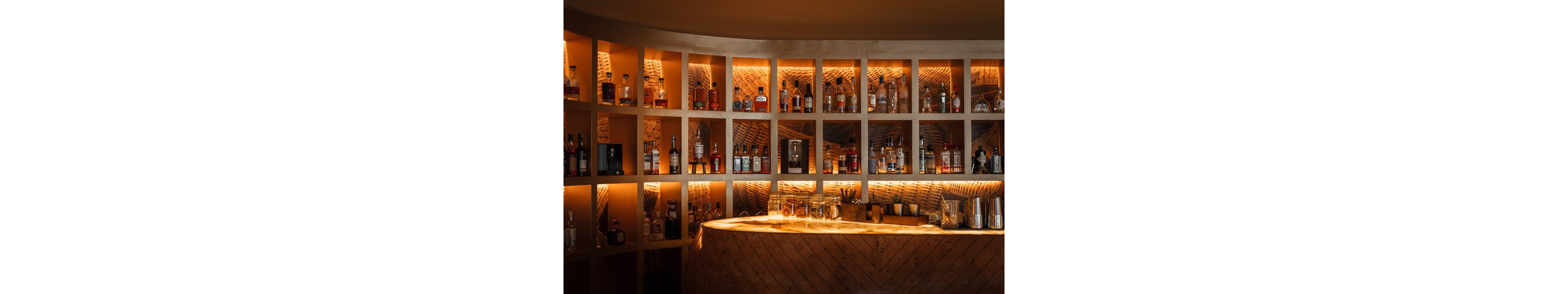 A stylish bar interior features a curved wooden counter in front of backlit shelves filled with assorted liquor bottles. The shelves are illuminated with warm amber lighting, highlighting the glassware and spirits on display. Metal shakers, bar tools, and glasses sit on the counter, suggesting a ready-to-serve cocktail setting. The overall atmosphere feels cozy and upscale, with rich wood textures and soft, glowing light.