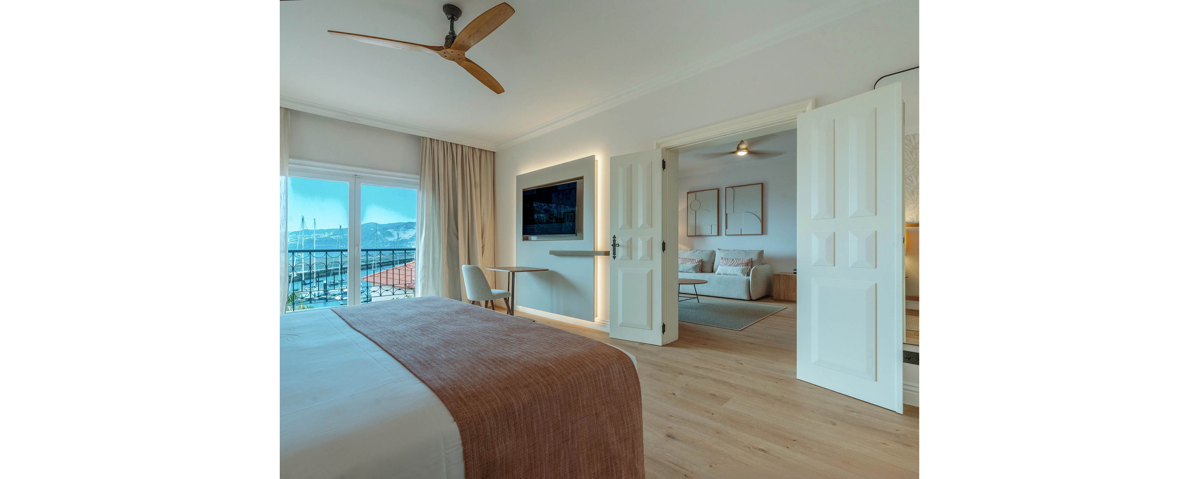 A modern hotel bedroom features a neatly made bed with a soft brown throw and light wood flooring. Large glass doors open to a balcony with a coastal view of mountains and rooftops. A wall-mounted television and small desk sit opposite the bed, while double doors lead to a cozy lounge area with sofa and artwork. Natural light and neutral tones create a calm, inviting atmosphere throughout the space.
