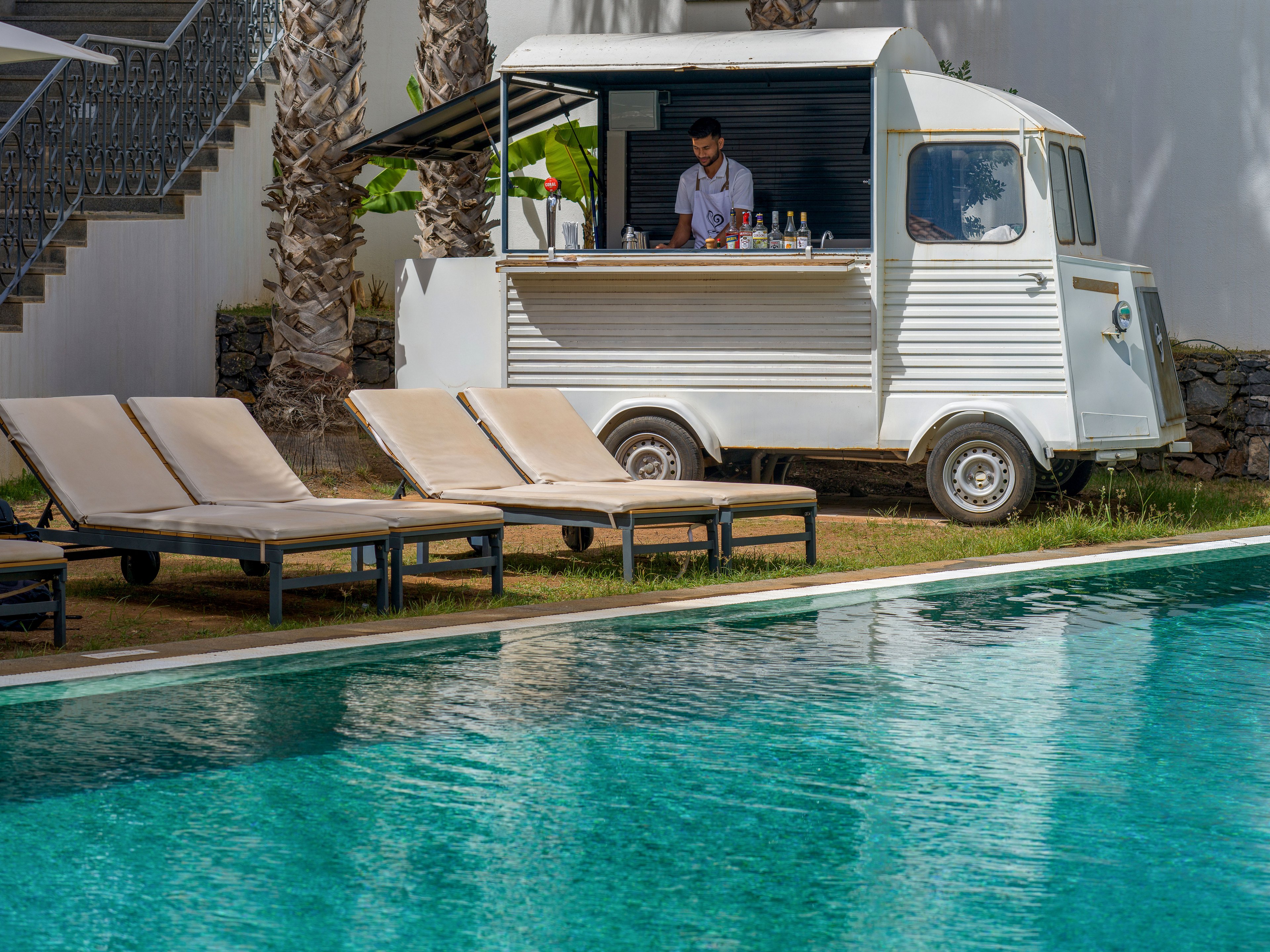 A mobile bar truck is parked beside a clear turquoise swimming pool. A bartender stands inside the retro-style vehicle preparing drinks. Several empty lounge chairs line the poolside area under partial shade from nearby palm trees. The scene suggests a relaxed resort environment with calm, sunny weather.