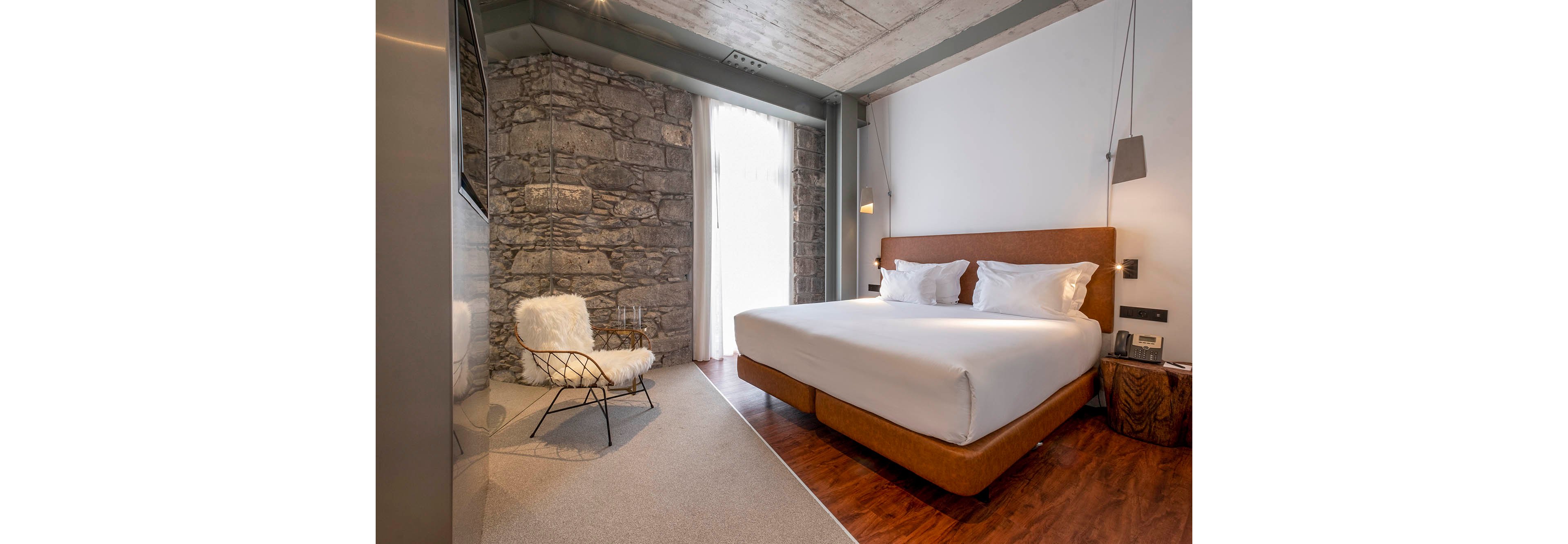 Contemporary hotel-style bedroom featuring a large double bed with white linens and plush pillows. The room combines a rustic stone accent wall with sleek modern finishes and warm wooden flooring. A minimalist metal chair with a fluffy white cushion sits near the window, which is covered by sheer curtains letting in soft natural light. Neutral tones and clean lines create a calm, inviting atmosphere suitable for relaxation or travel stays.